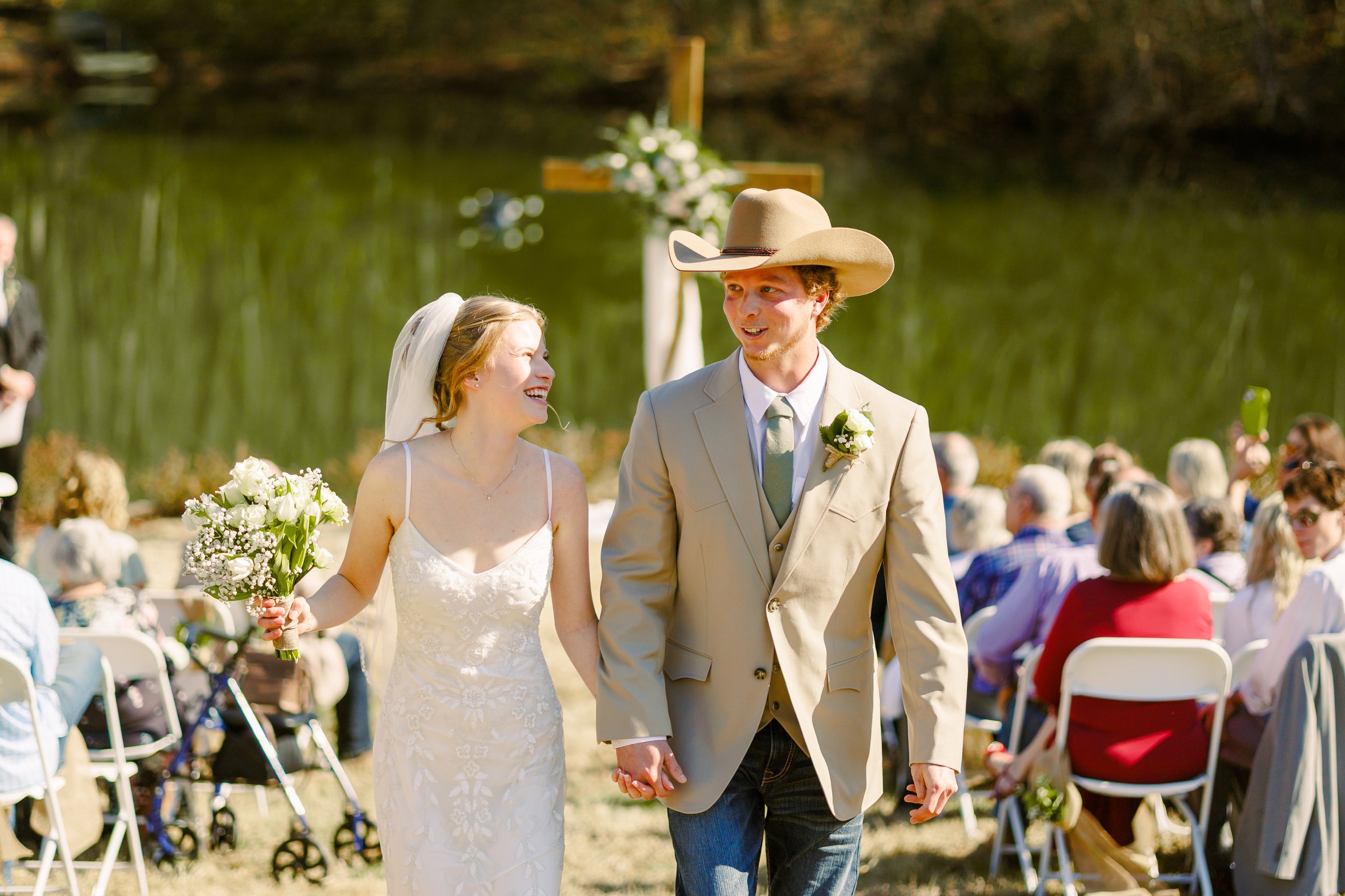 A bride and groom holding hands during their outdoor wedding ceremony. The bride is smiling and holding a bouquet of flowers, while the groom is wearing a cowboy hat and a light gray suit jacket. In the background, there is a wooden cross decorated w