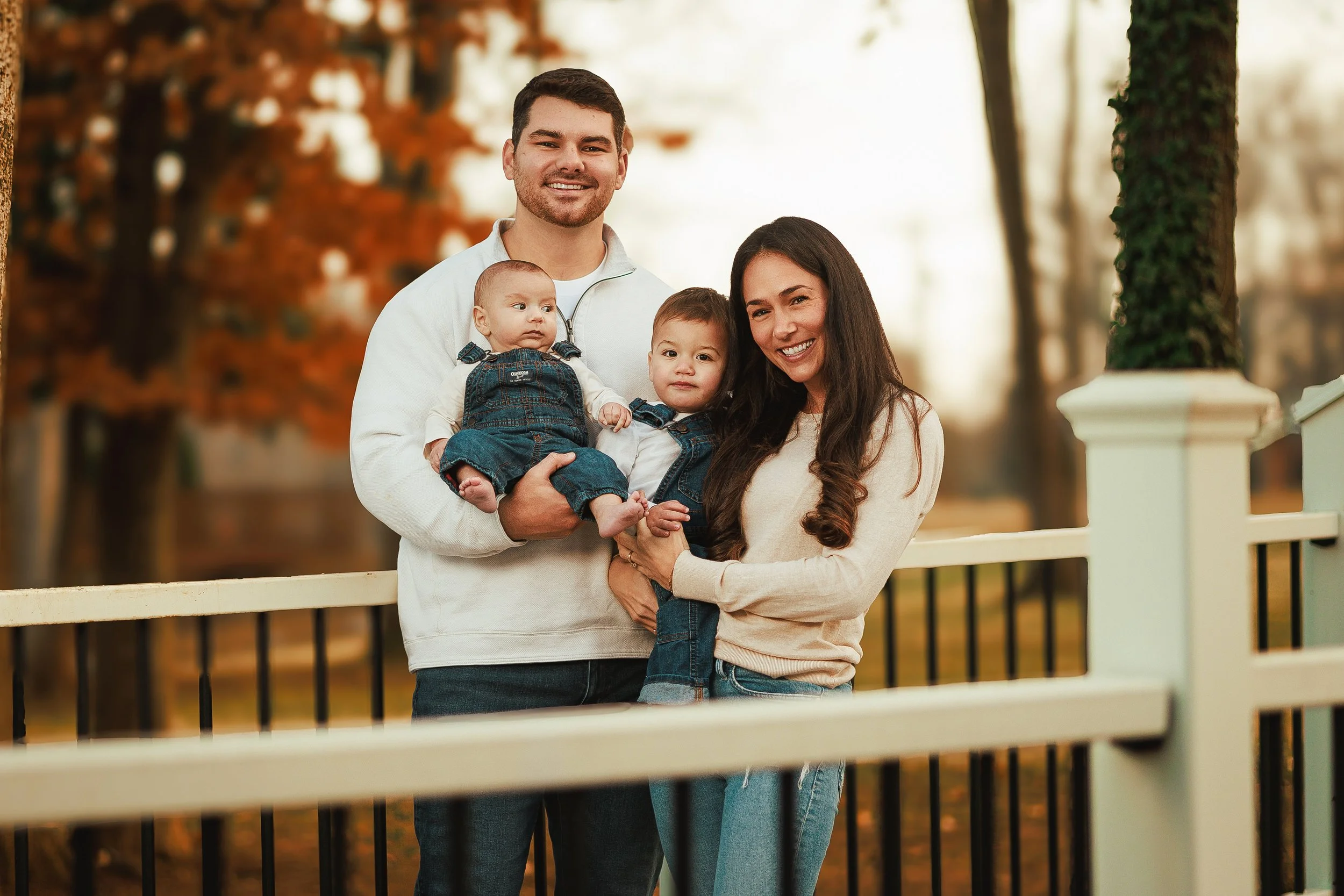 A happy family outdoor in autumn on a walkway near trees with orange leaves, posing together for a photo. The father holds a baby, and a young girl stands with her mother.