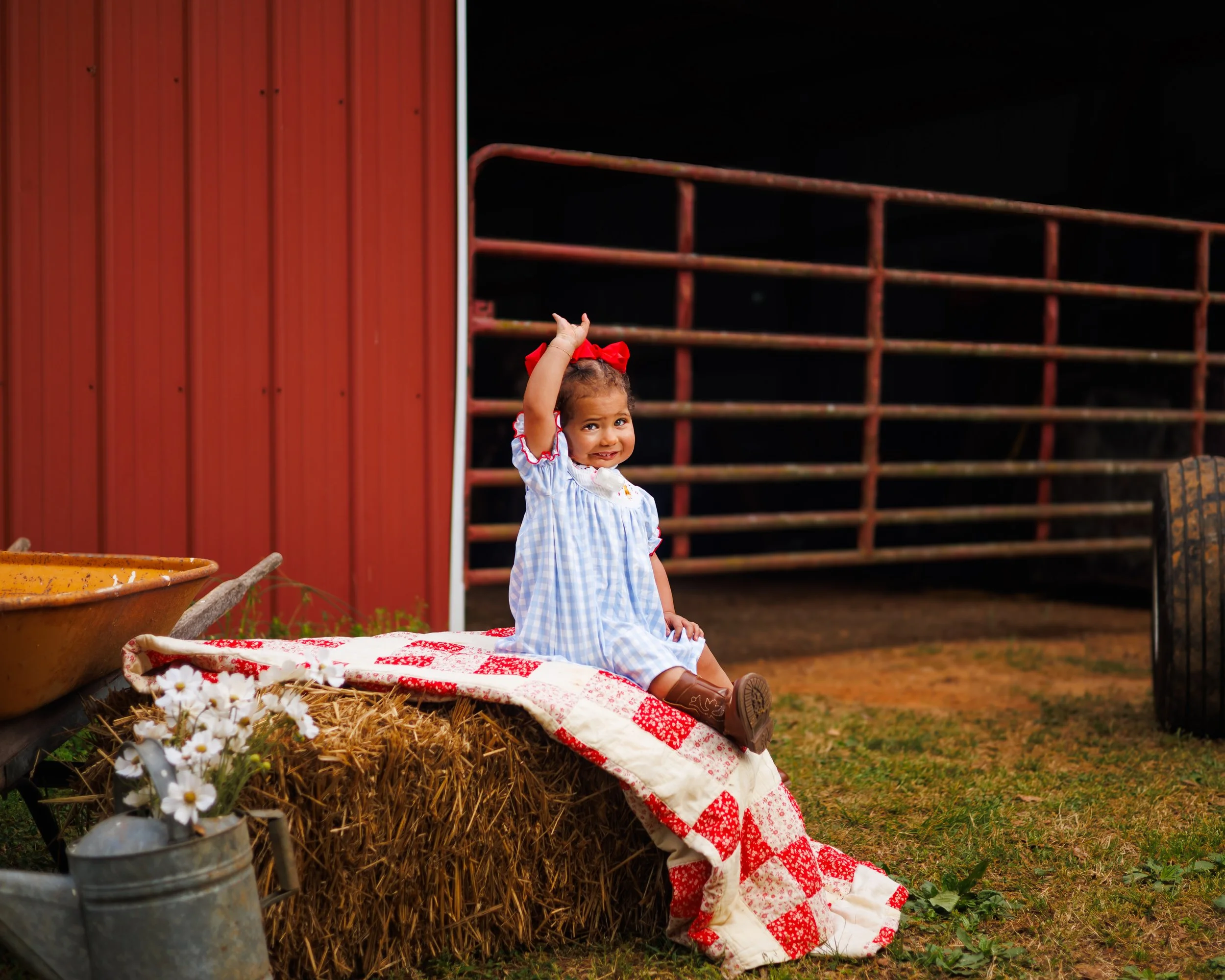 A young girl with curly hair tied with a red bow, sitting on a hay bale, waving, wearing a blue and white checkered dress with red trim, in front of a red barn with a metal fence and farm tools.