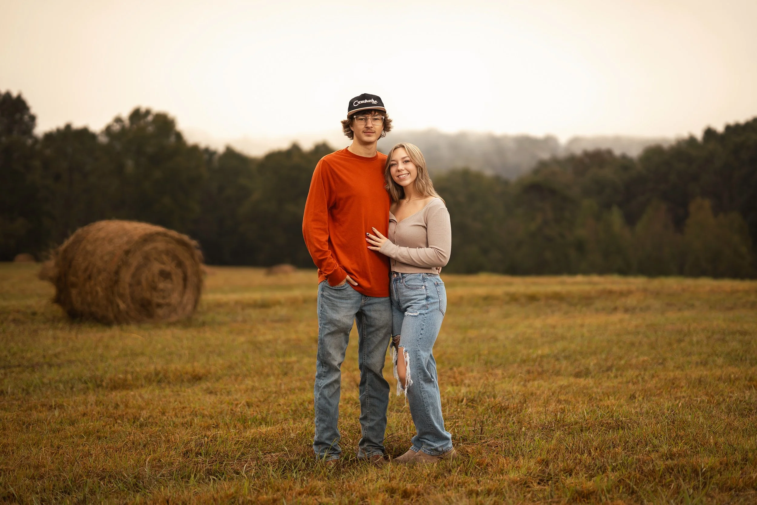 A young man and woman standing together in a field with hay bales, with trees and a soft sky in the background.