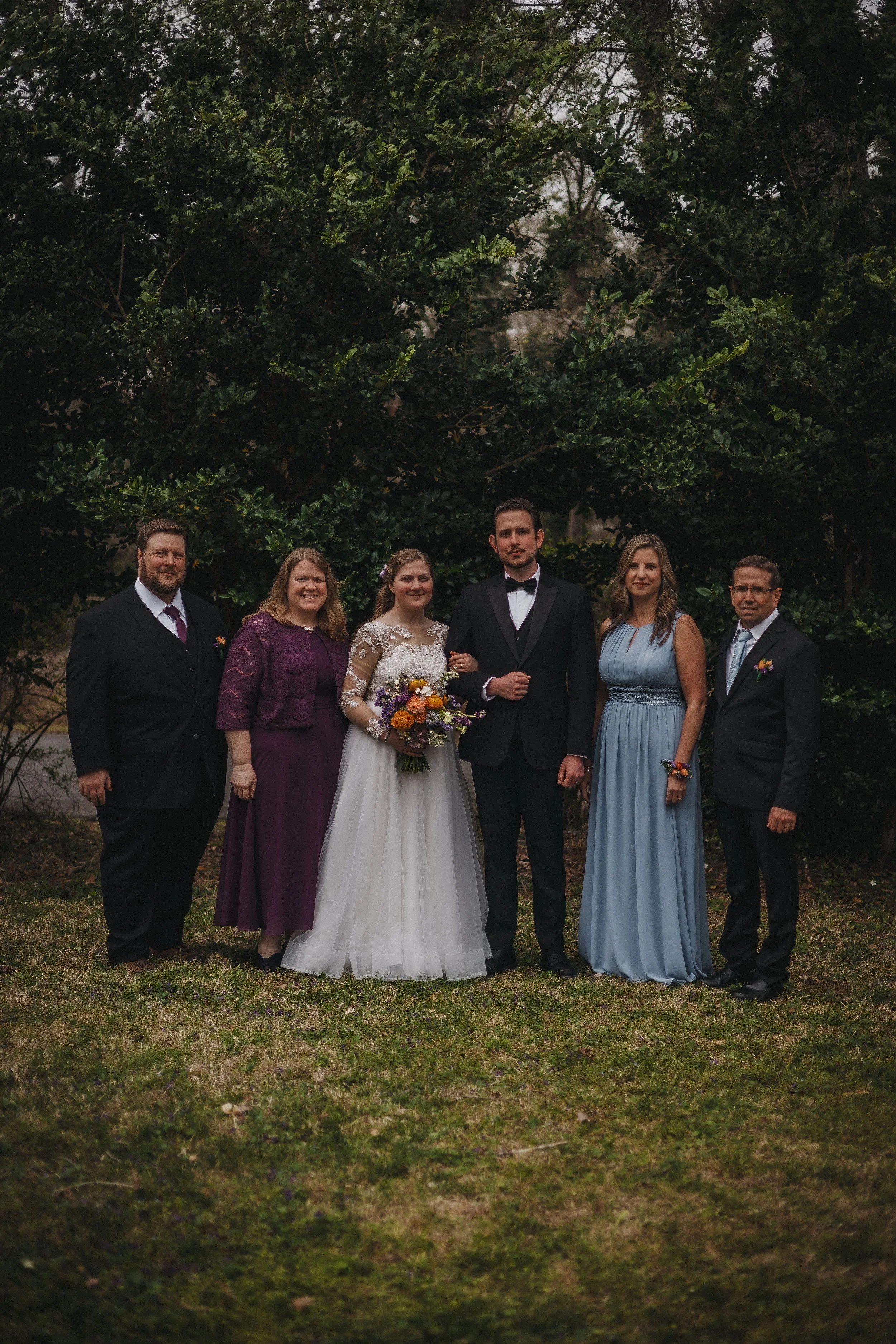 A group of seven people, including a bride and groom, standing outdoors on grass with trees in the background