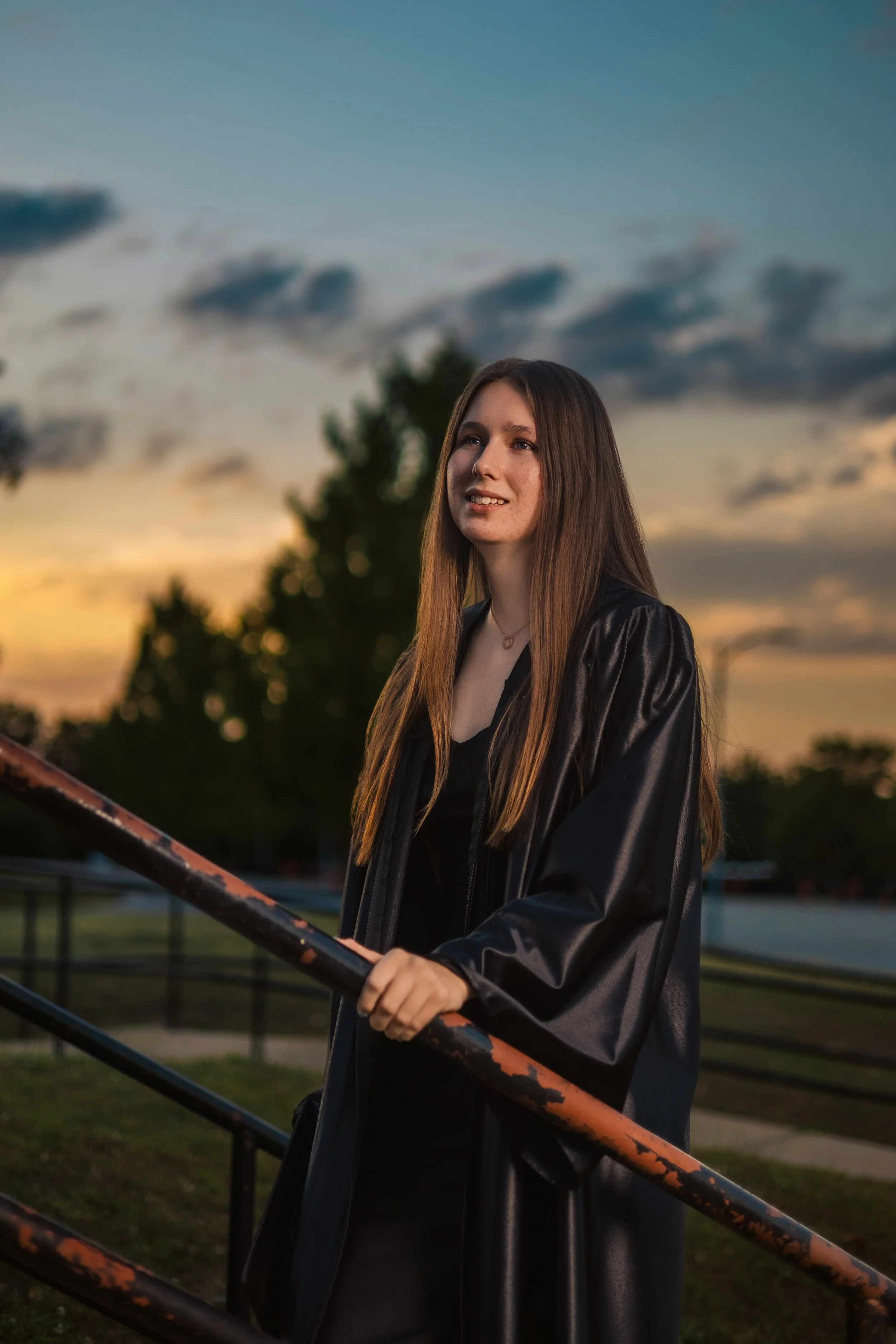 A young woman with long brown hair wearing a black leather jacket stands outdoors at sunset, holding onto a metal railing, with a background of trees and a cloudy sky.