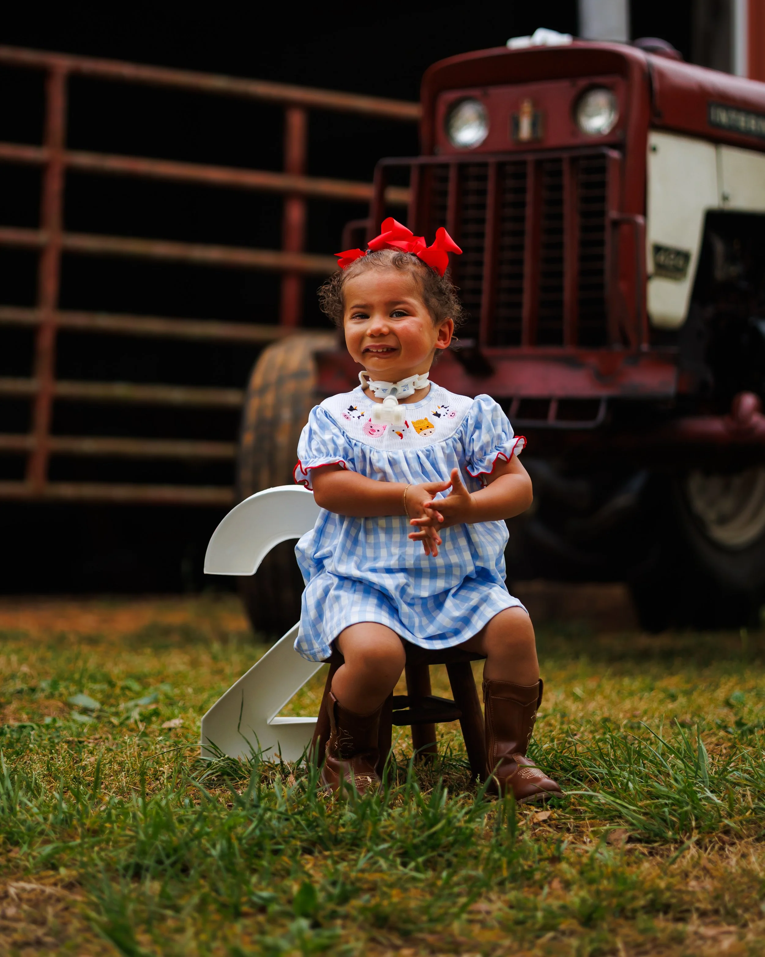 A young girl with curly hair, red bow, and a blue and white checkered dress sitting on a small stool in a yard with grass, with a tractor in the background.