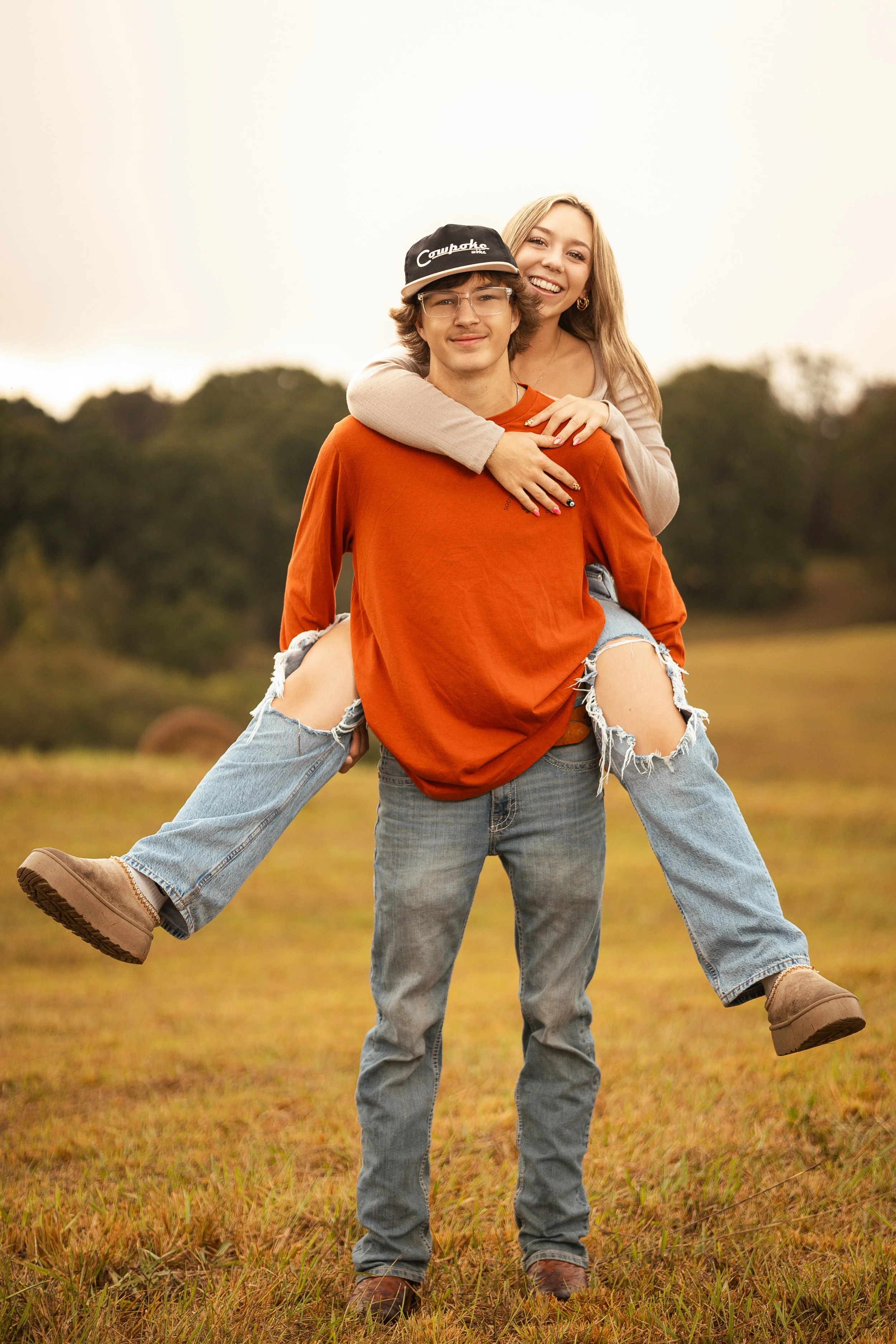 A young man in a red long-sleeve shirt with glasses and a black cap carrying a young woman with blonde hair on his back in a grassy field with trees in the background.