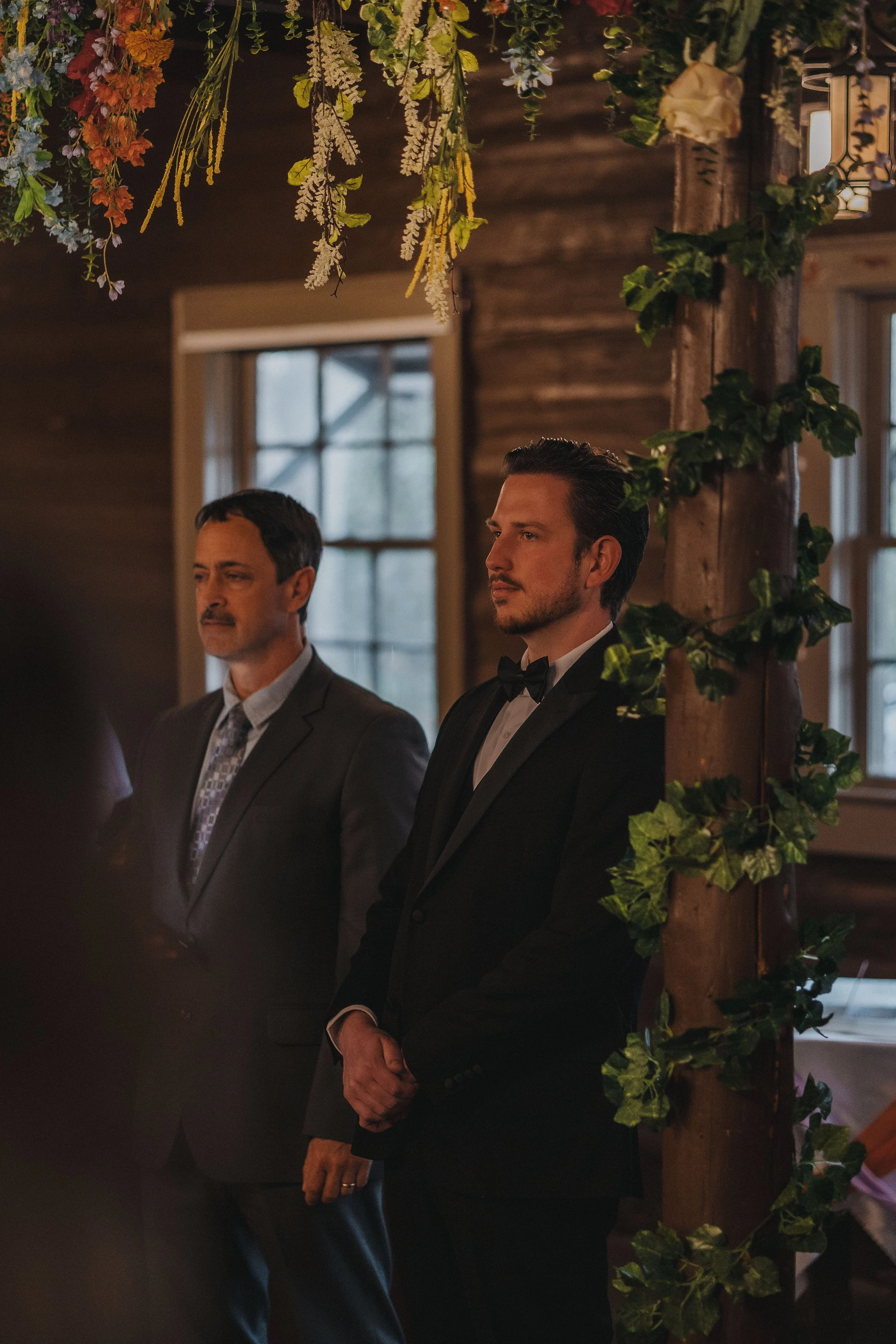 Two men in formal attire standing indoors near a wooden pillar decorated with green ivy and hanging flowers, with large windows in the background.