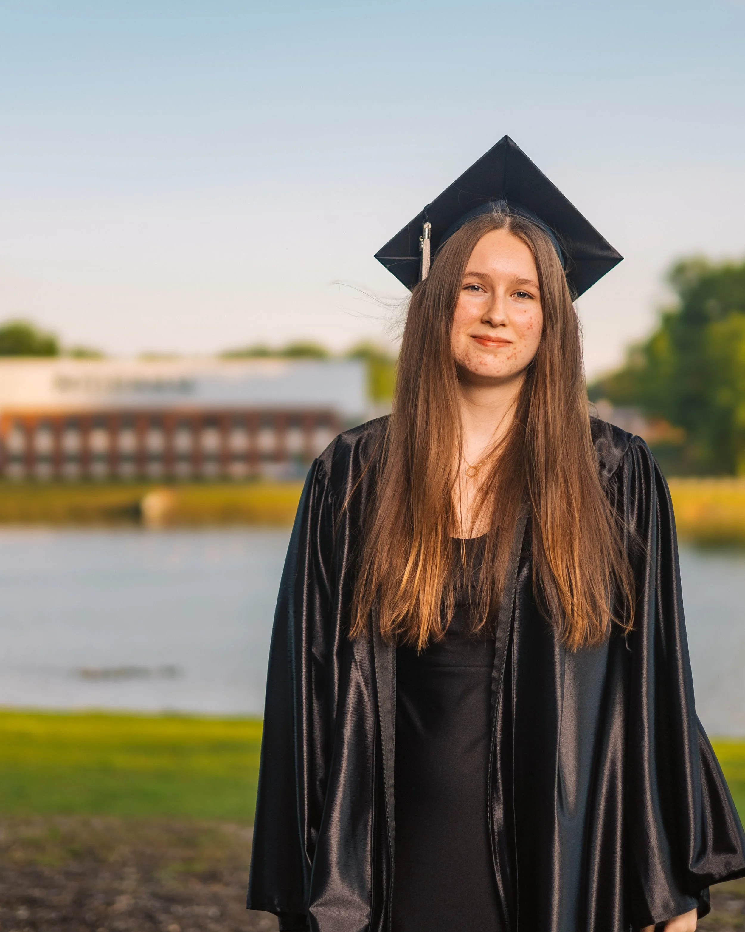 A young woman in a black graduation gown and cap standing outdoors near a lake or river, with trees and a building in the background during daylight.