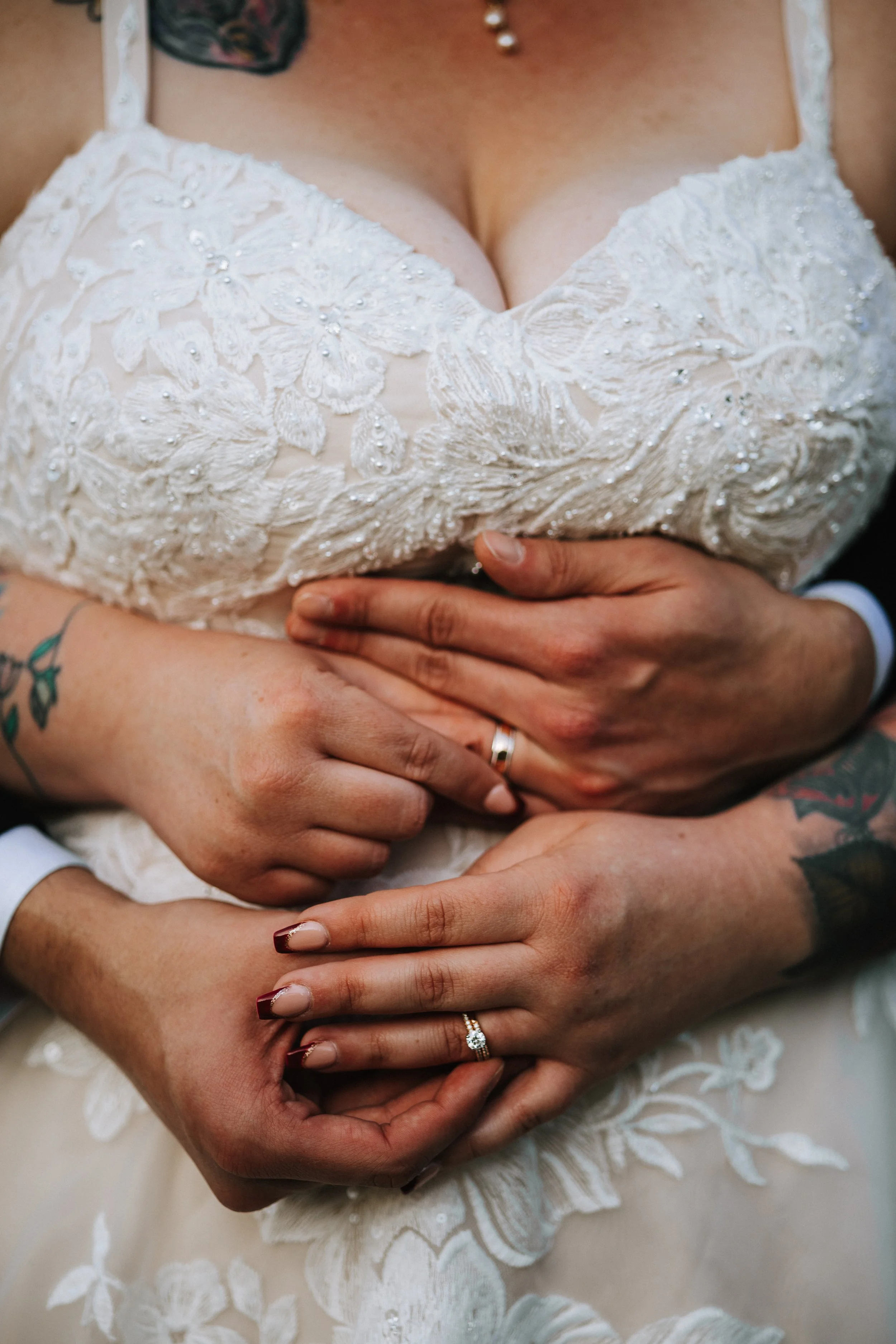 Close-up of a bride in a lace wedding dress with a deep neckline, featuring detailed embroidery, beads, and pearls. Multiple hands, possibly her or her close family and friends, are gently holding her waist and hands, some with rings visible, includi