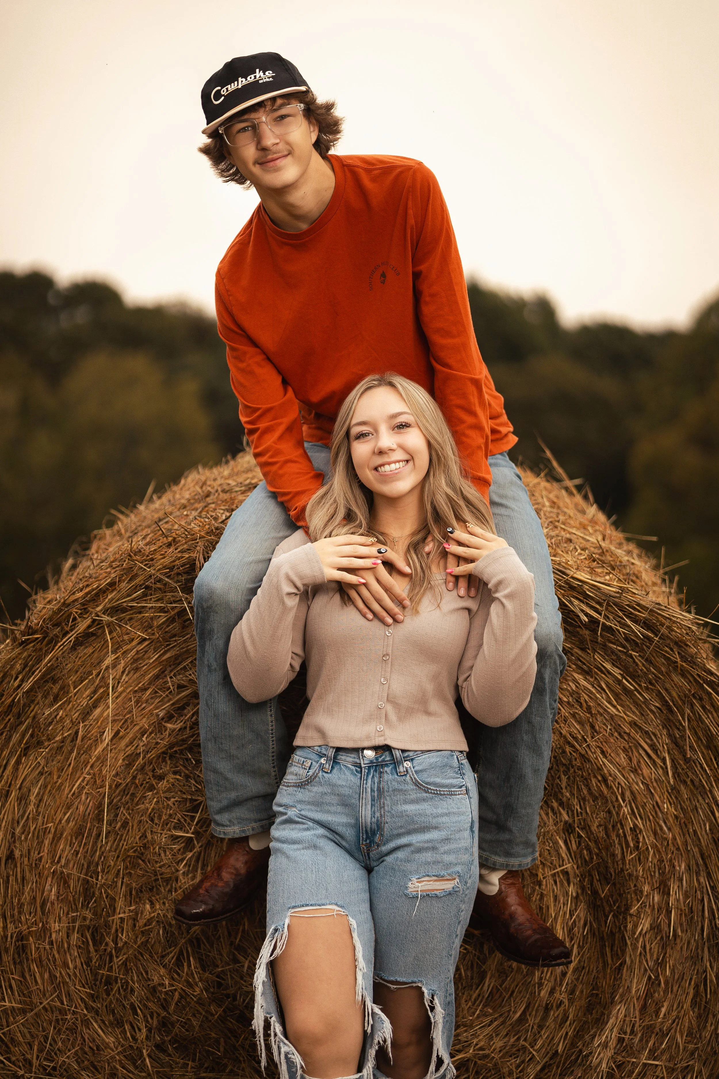 Three young adults sitting on a hay bale outdoors during sunset, smiling and enjoying each other's company.
