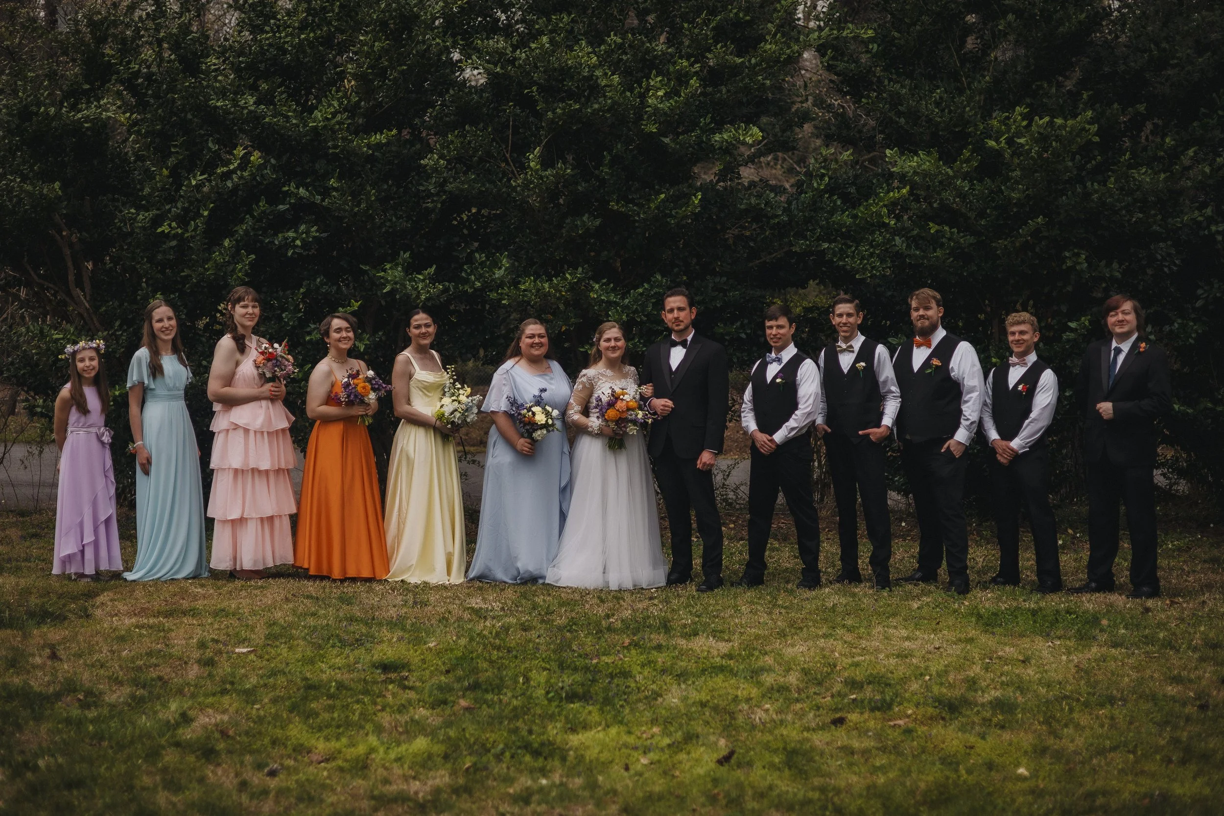 A group of people dressed in formal attire, including women in colorful dresses and men in suits and vests, standing outdoors on grass with trees in the background, for a wedding or celebration.