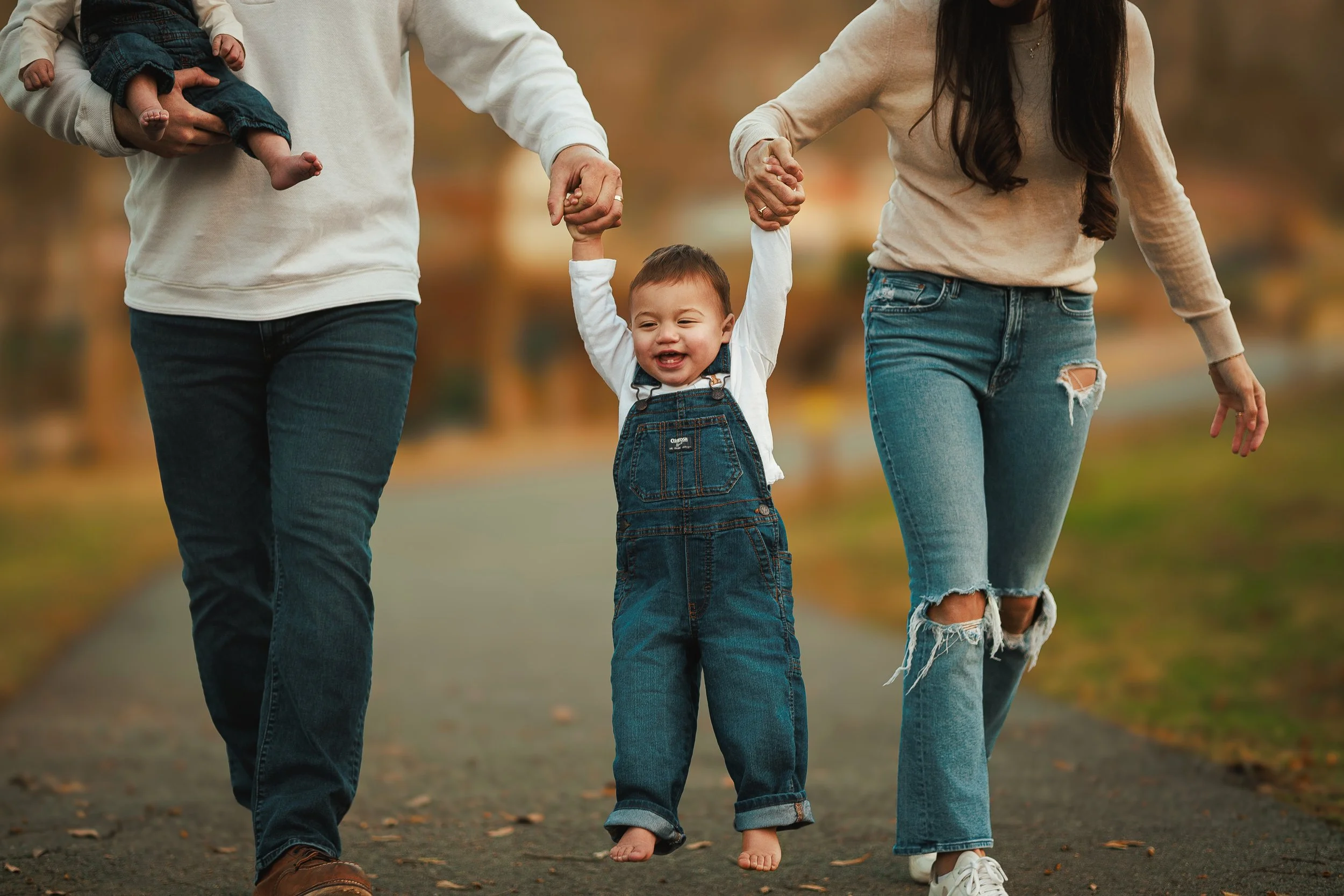 A family walking outdoors in a park during autumn, holding a young child's hands up as the child walks barefoot on the ground.