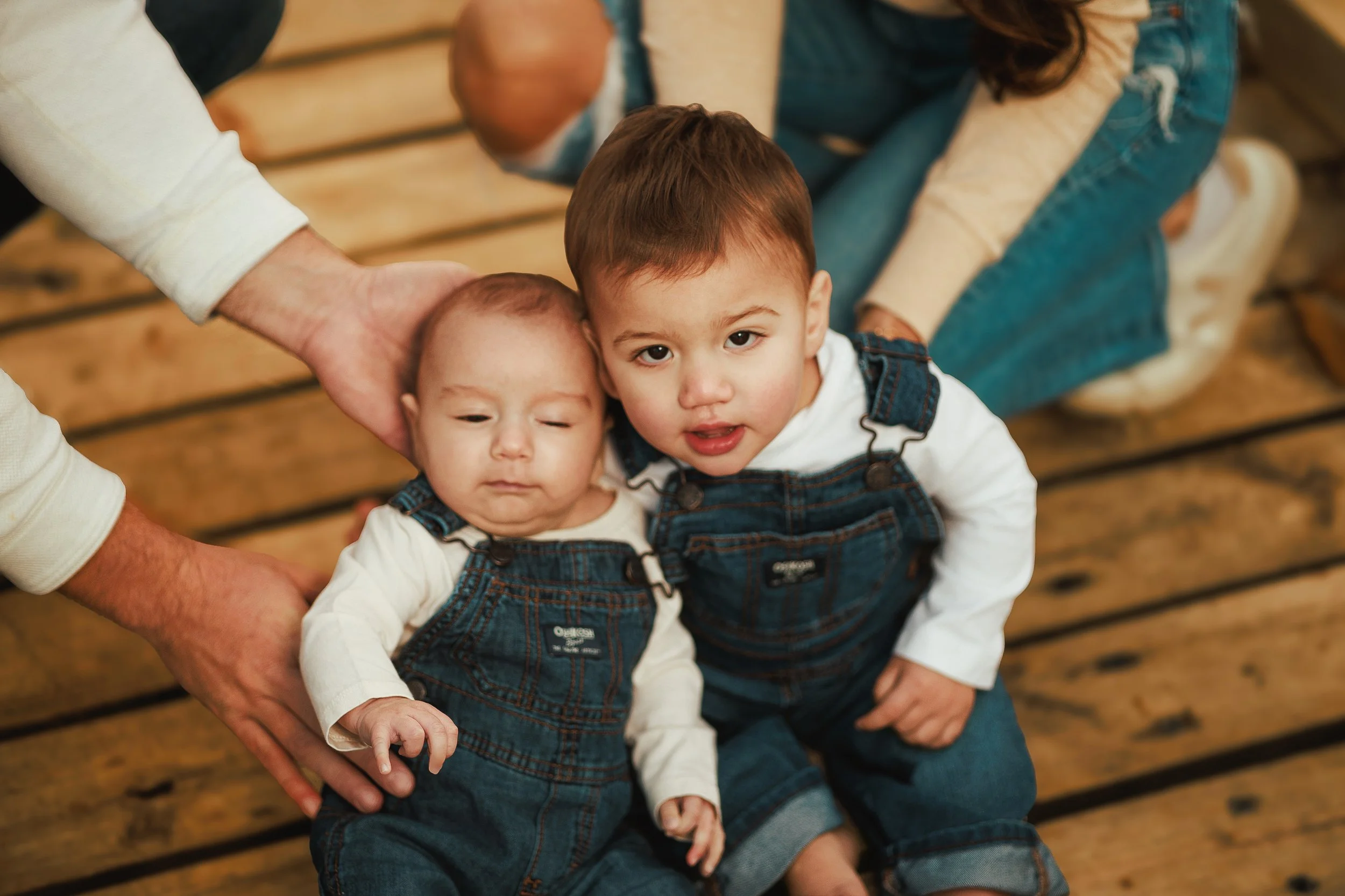 Two young boys dressed in denim overalls sitting on a wooden bench, being held and supported by adults. The children have light skin and brown hair, and they are looking at the camera.