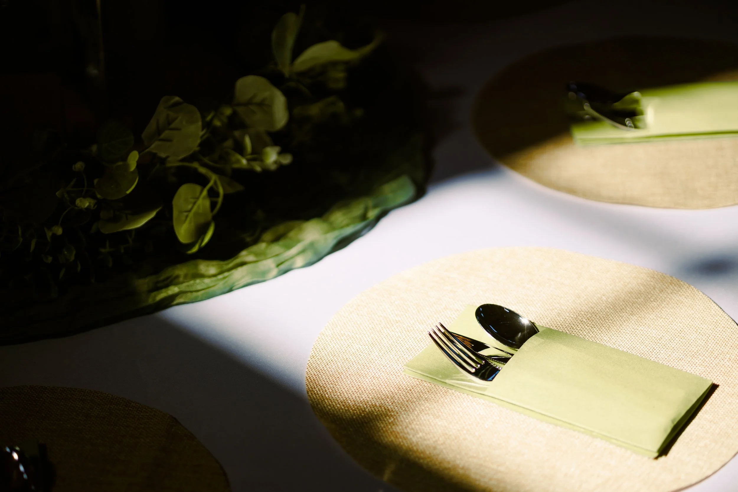 Part of a table setting with a fork and spoon on a yellow napkin, and a place mat under the napkin. In the background, there is a dark, textured bowl with green leaves, and some abstract objects with light and shadow.