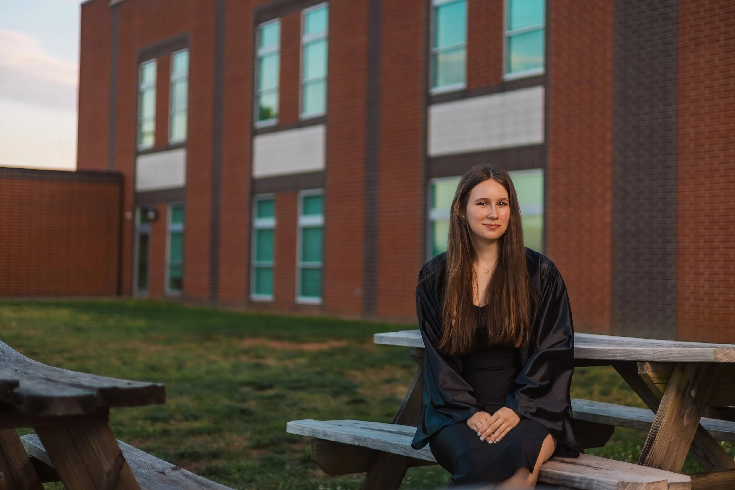 A young woman with long brown hair sitting on a wooden picnic table outside a brick building with large windows, during what appears to be sunset or dusk.
