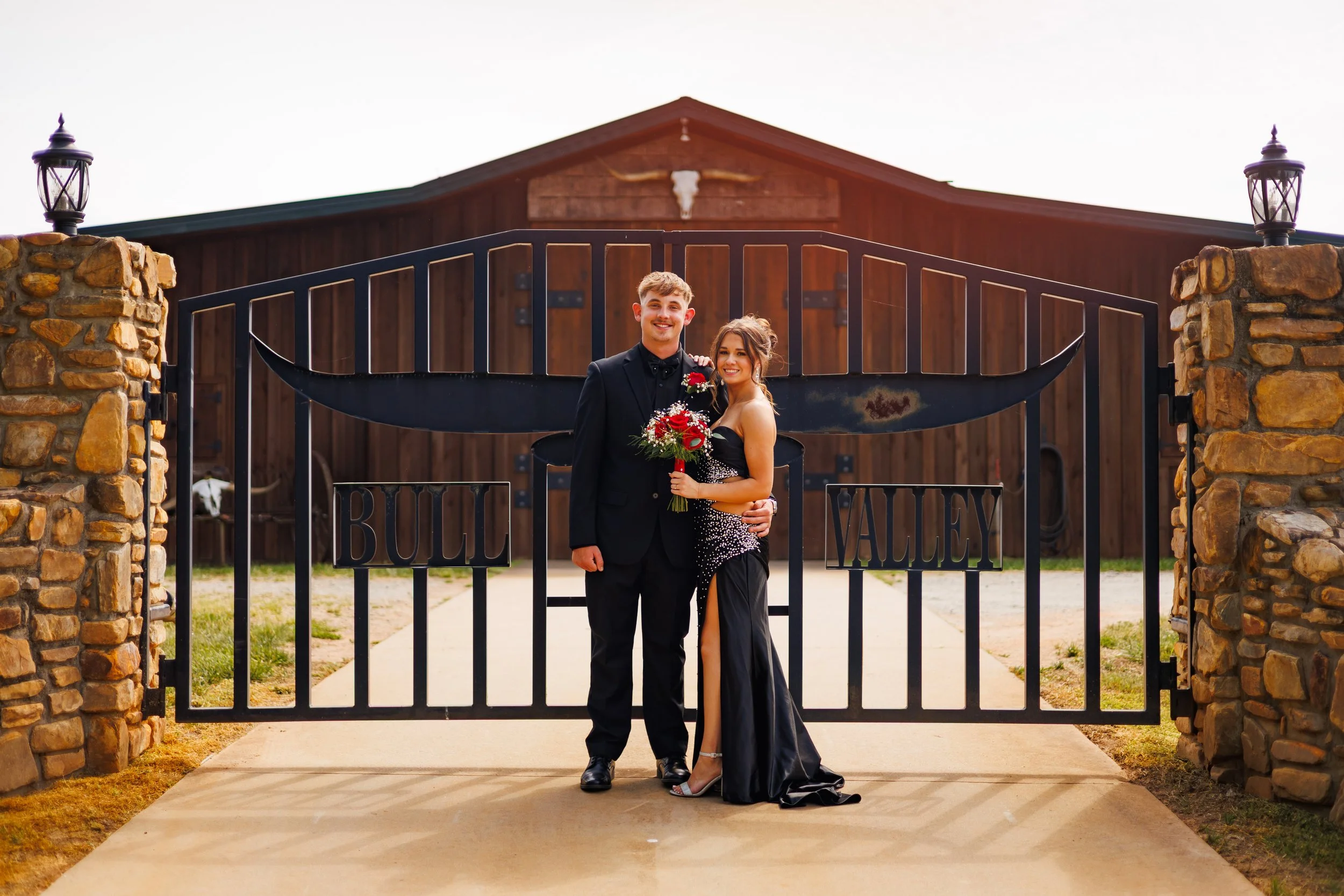 A smiling young man and woman dressed in formal attire standing in front of a gate with a sign that says 'Bullet Valley,' with a barn-like building and stone pillars in the background.