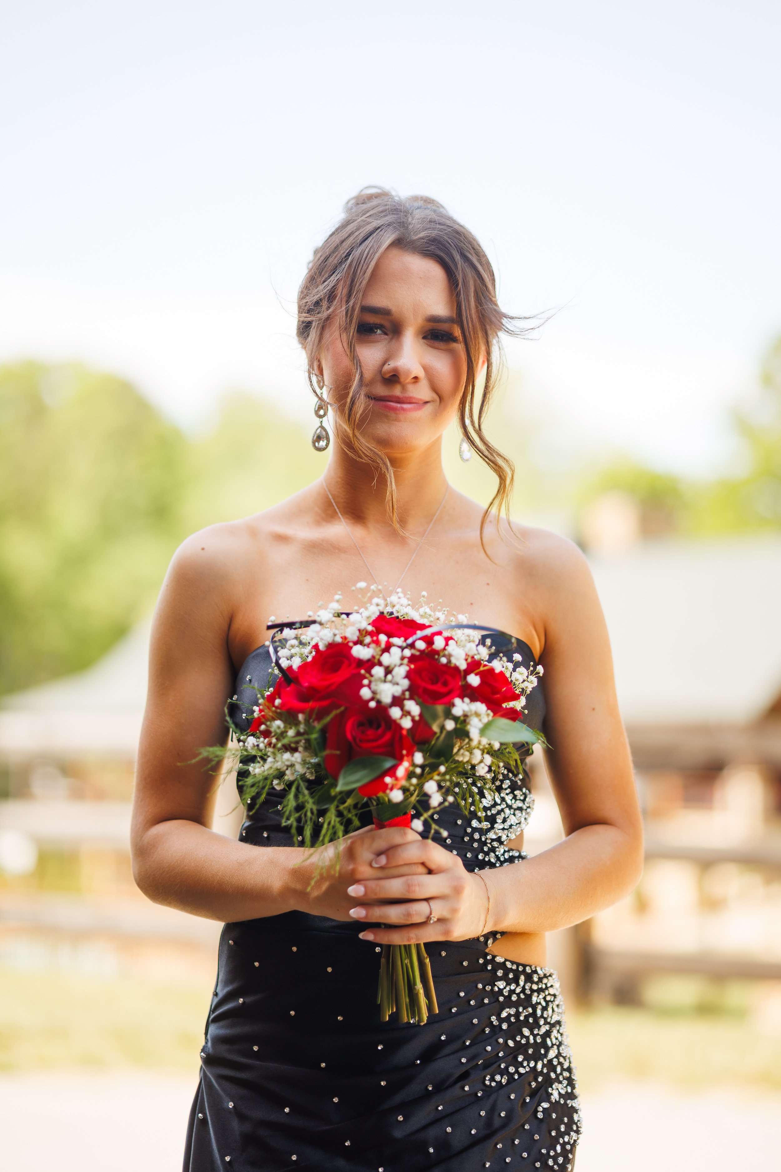 A woman in a black dress with silver embellishments holding a bouquet of red roses and white baby's breath flowers, outdoors with blurred trees in the background.