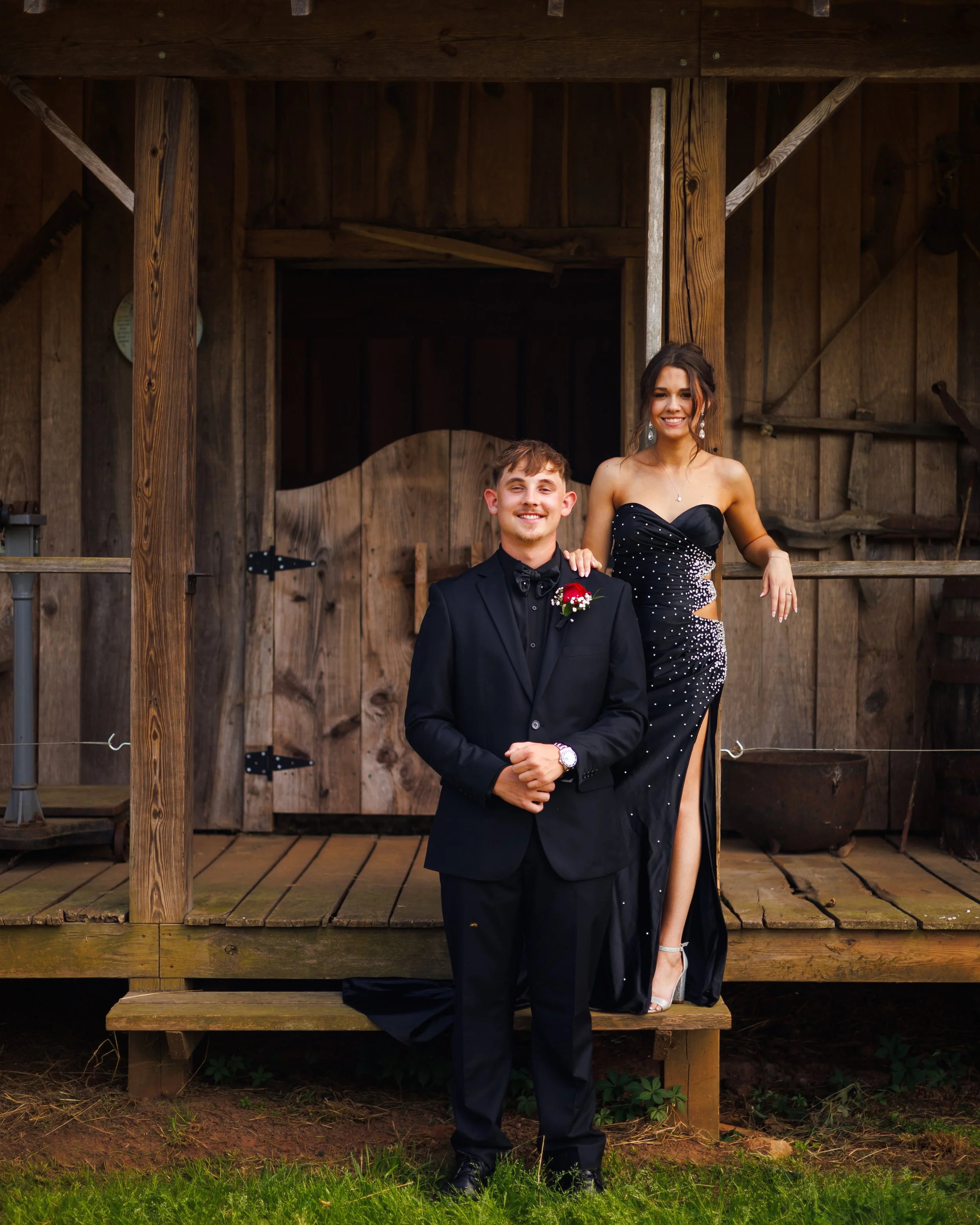 A couple dressed in formal attire standing in front of a rustic wooden barn. The man is wearing a black tuxedo with a bow tie and a red boutonniere, and the woman is wearing a black strapless gown with sequins and a thigh-high slit, smiling at the ca
