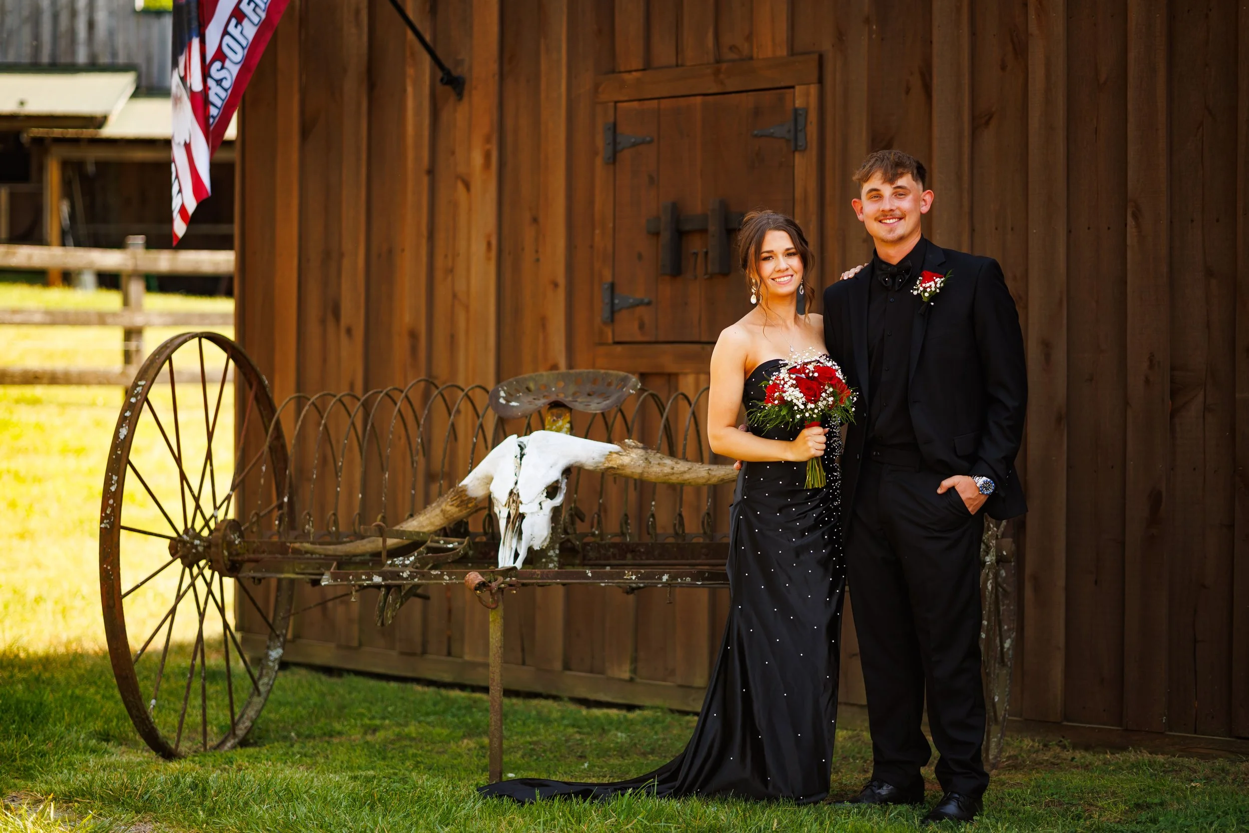 A young couple dressed in formal attire, standing outdoors against a wooden background, smiling. The woman is wearing a black dress with white polka dots and holding a bouquet of red and white flowers, while the man is in a black tuxedo with a bouton
