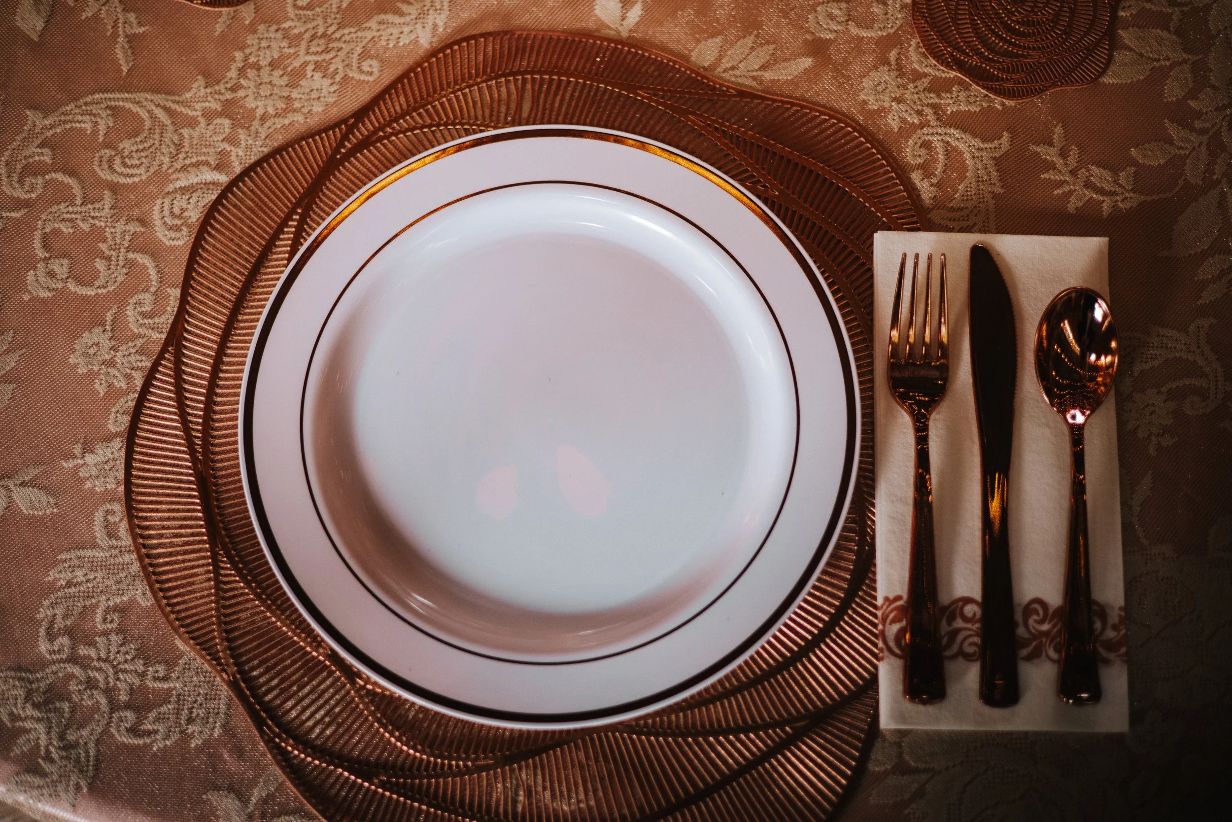 Empty white dinner plate with a black and gold rim, placed on a copper-colored wire placemat. Next to it, on a beige napkin, are a copper-colored fork, knife, and spoon. The tablecloth underneath has a floral and paisley pattern in shades of beige an
