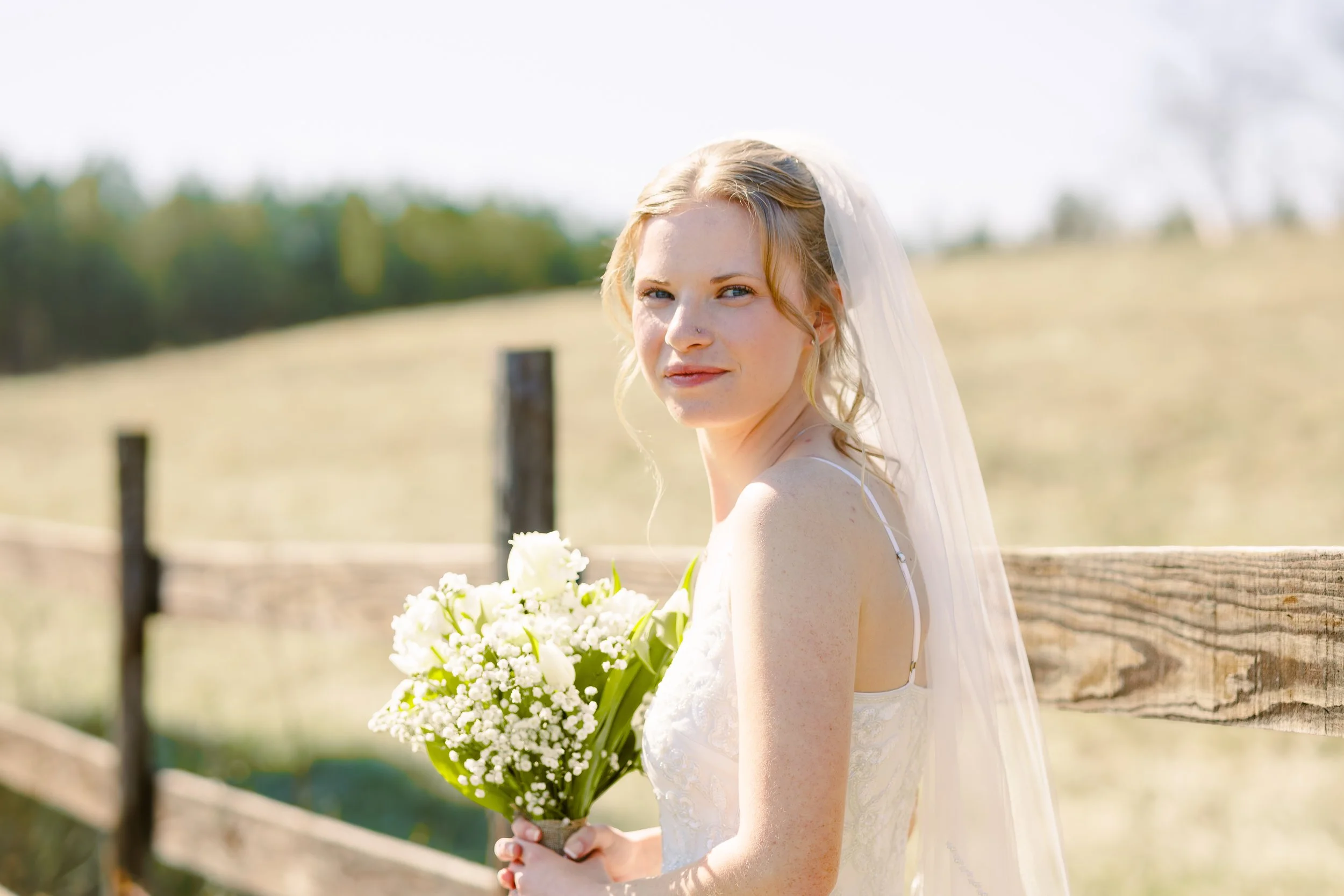 A bride with blonde hair wearing a white wedding dress and veil, holding a bouquet of white flowers, standing outdoors near a wooden fence with a blurry green landscape in the background.