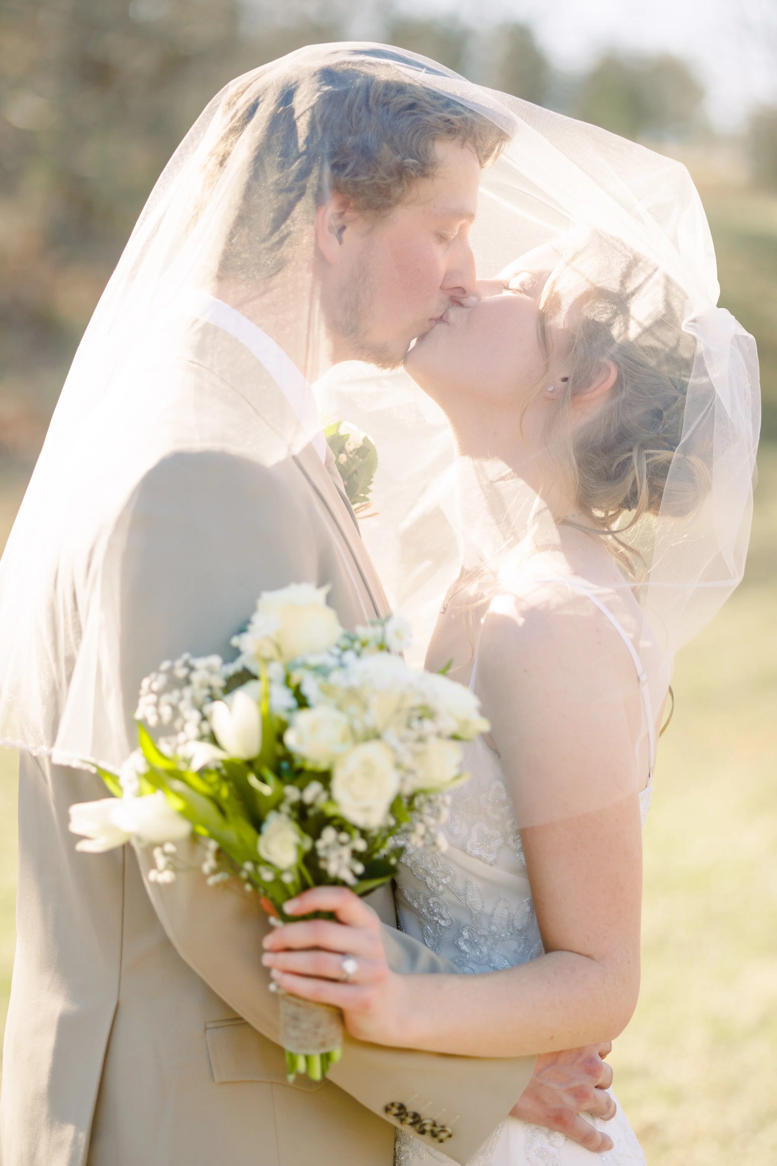 A bride and groom share a kiss under a sheer veil during their wedding outdoors, with soft sunlight illuminating the scene.