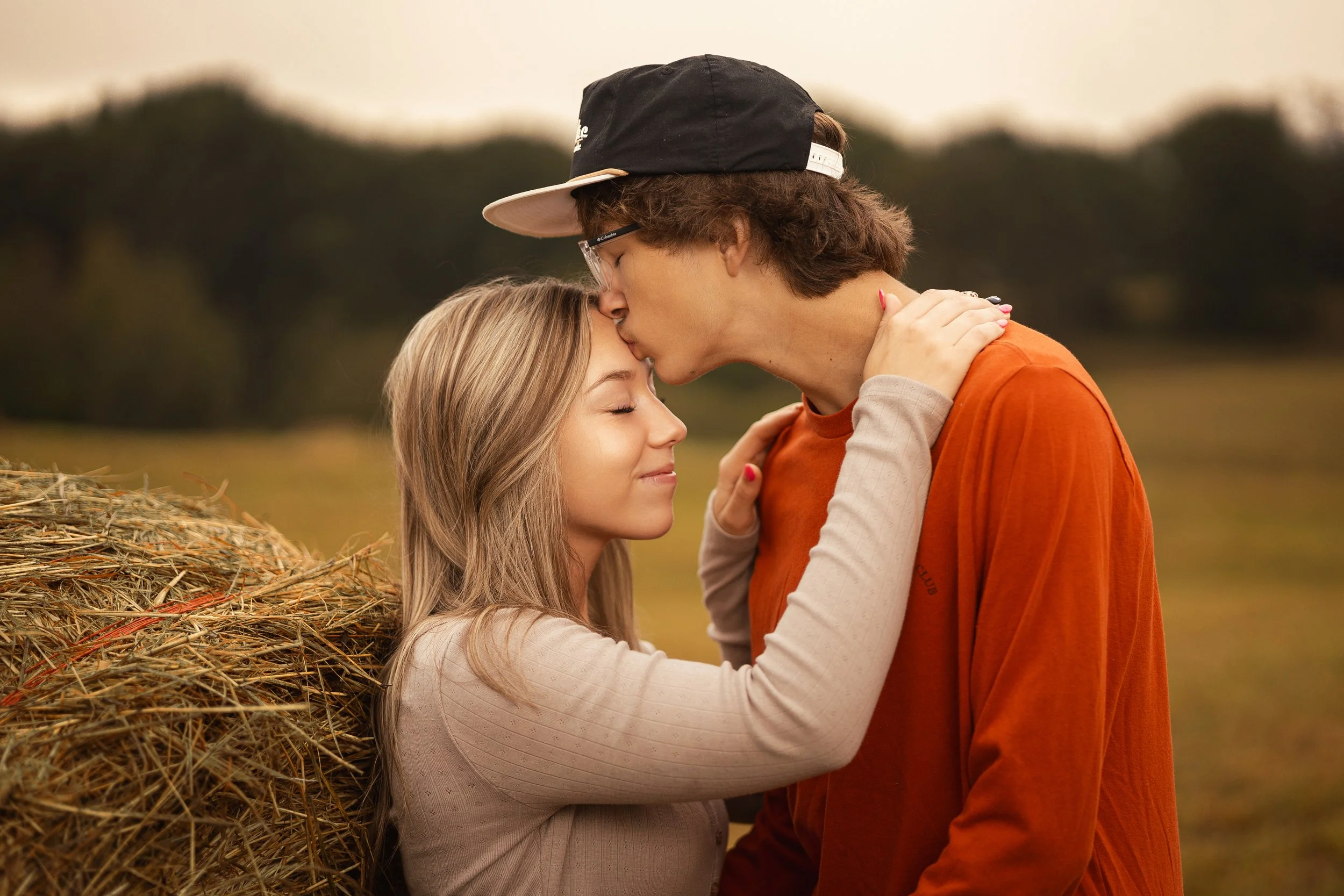 A young couple sharing a kiss with eyes closed, standing on a farm near a haystack during sunset.