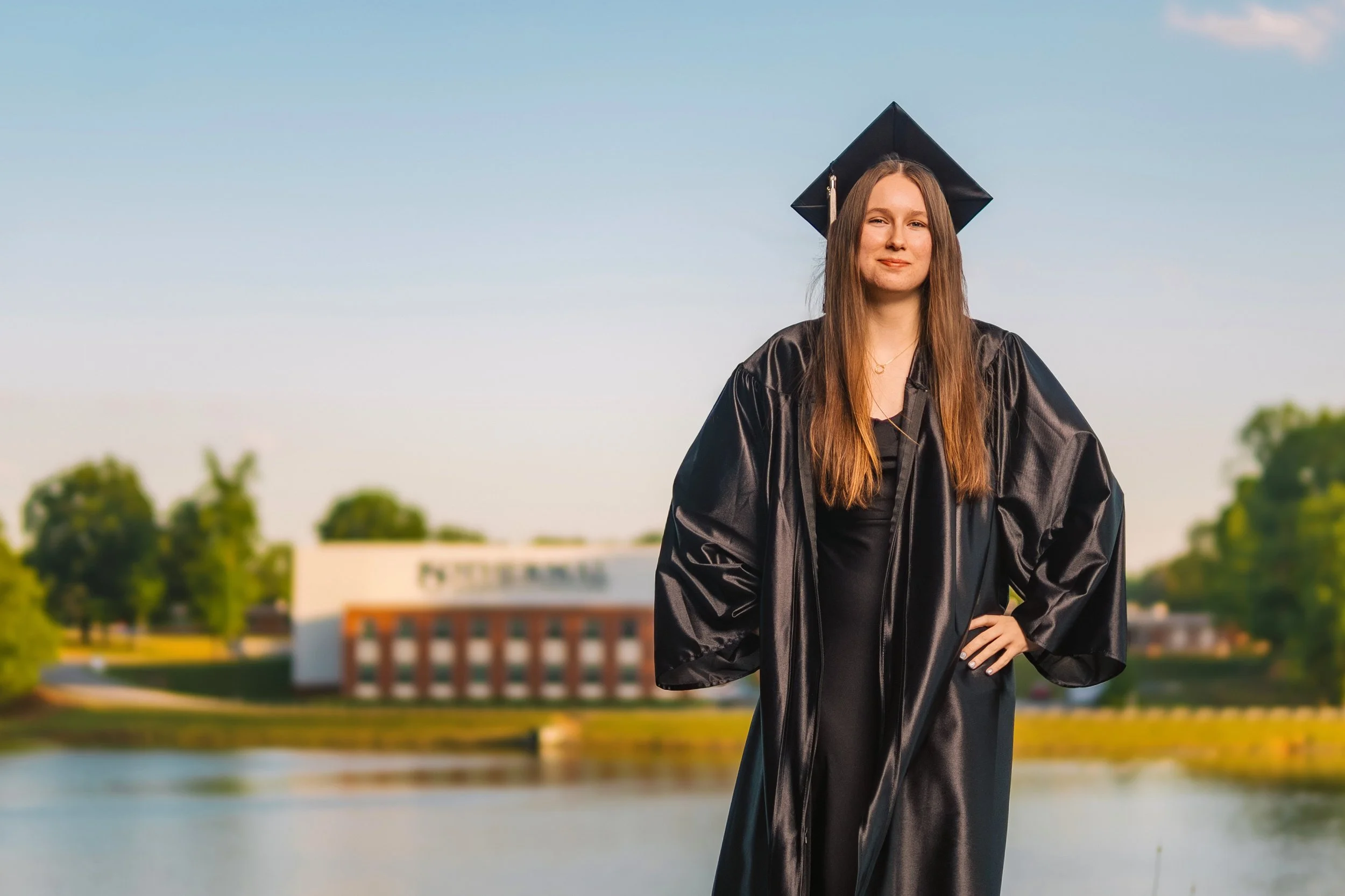 A young woman in a black graduation gown and cap standing outdoors near a river, with trees and a building in the background, during the daytime.