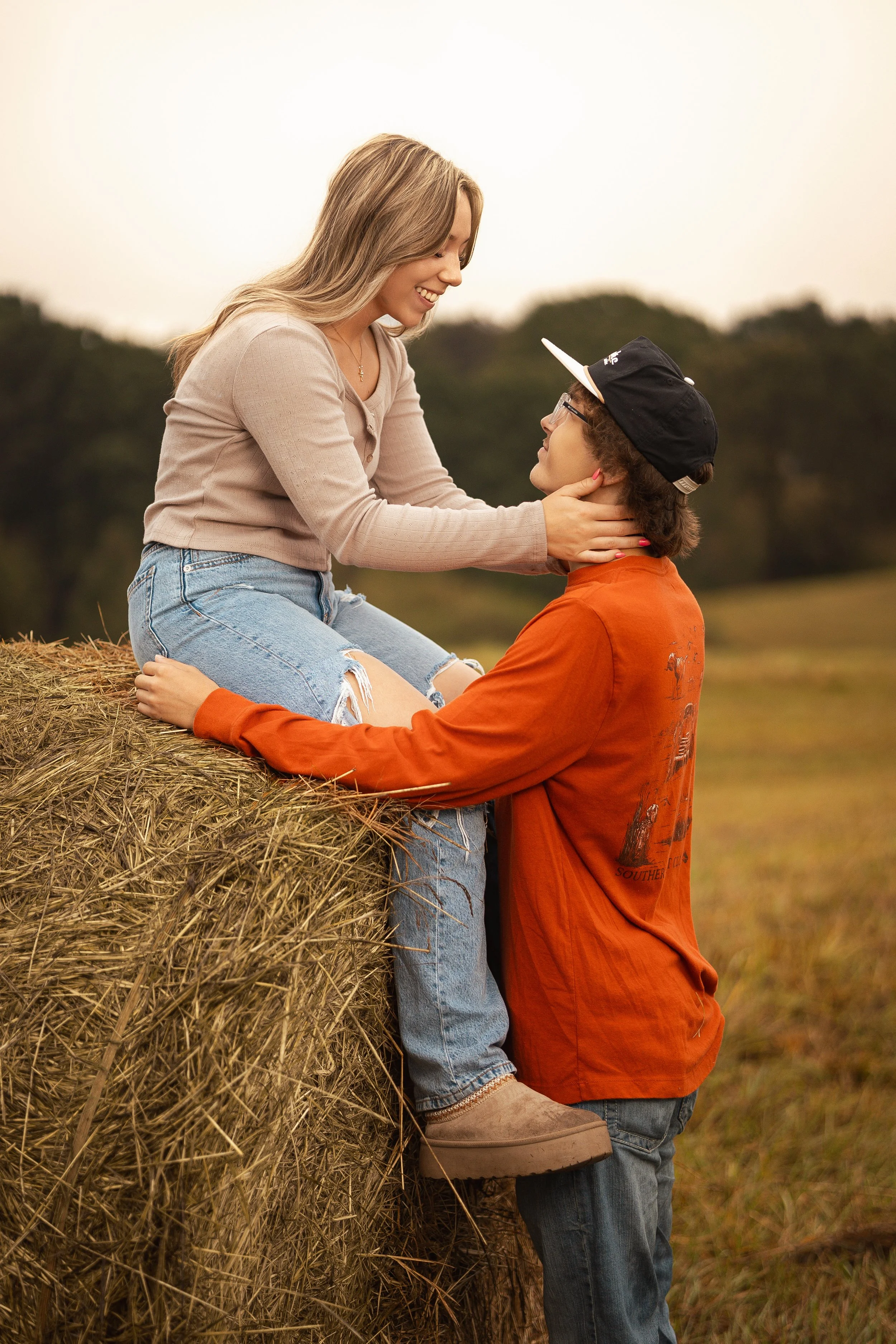 A woman with long blonde hair, wearing a beige shirt and ripped jeans, sitting on a hay bale, smiling at a young man with glasses and a backward cap, who is standing and holding her face affectionately, in a field during sunset.