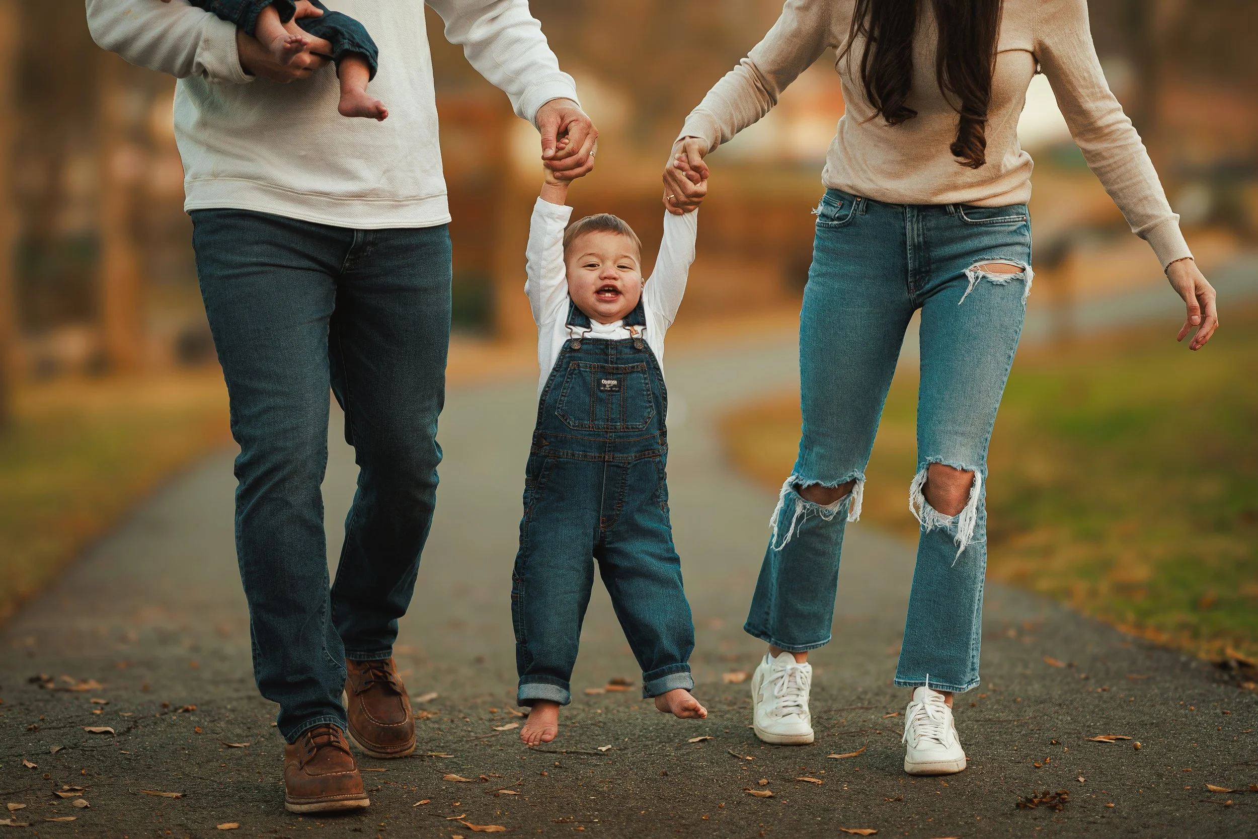 A family walking outdoors during autumn, with a child holding parents' hands, one parent holding a young child's hand, and the child's feet are barefoot while walking on a leaf-covered path.