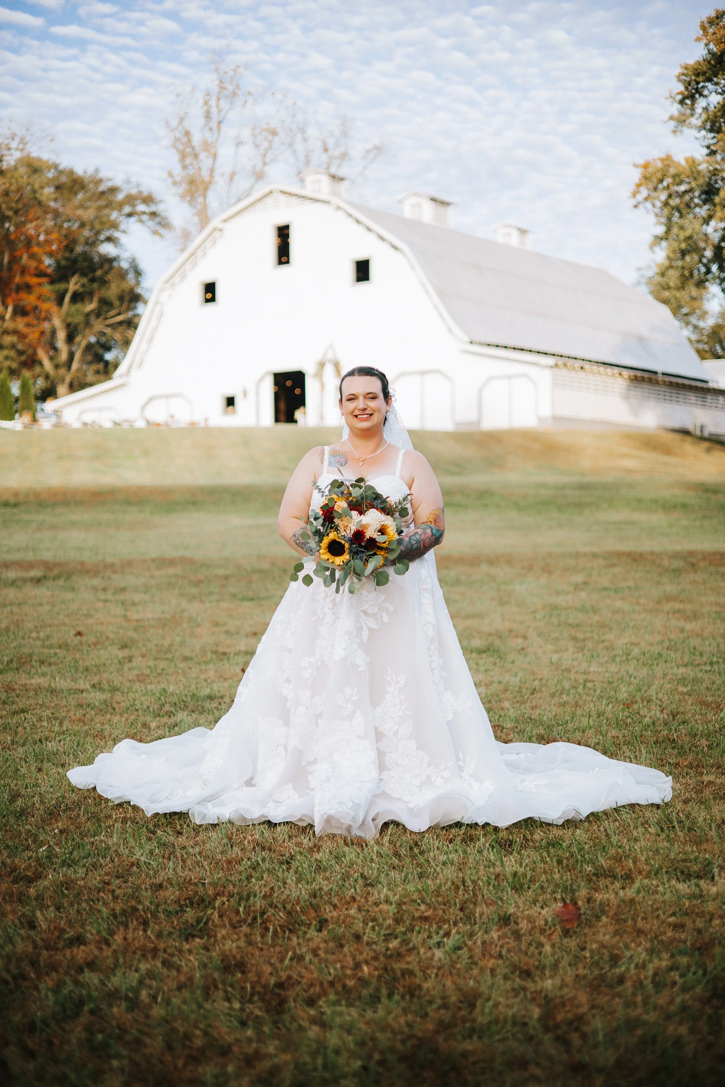 A bride in a white wedding gown standing on a grassy lawn, holding a bouquet of flowers, with a white barn and trees in the background.