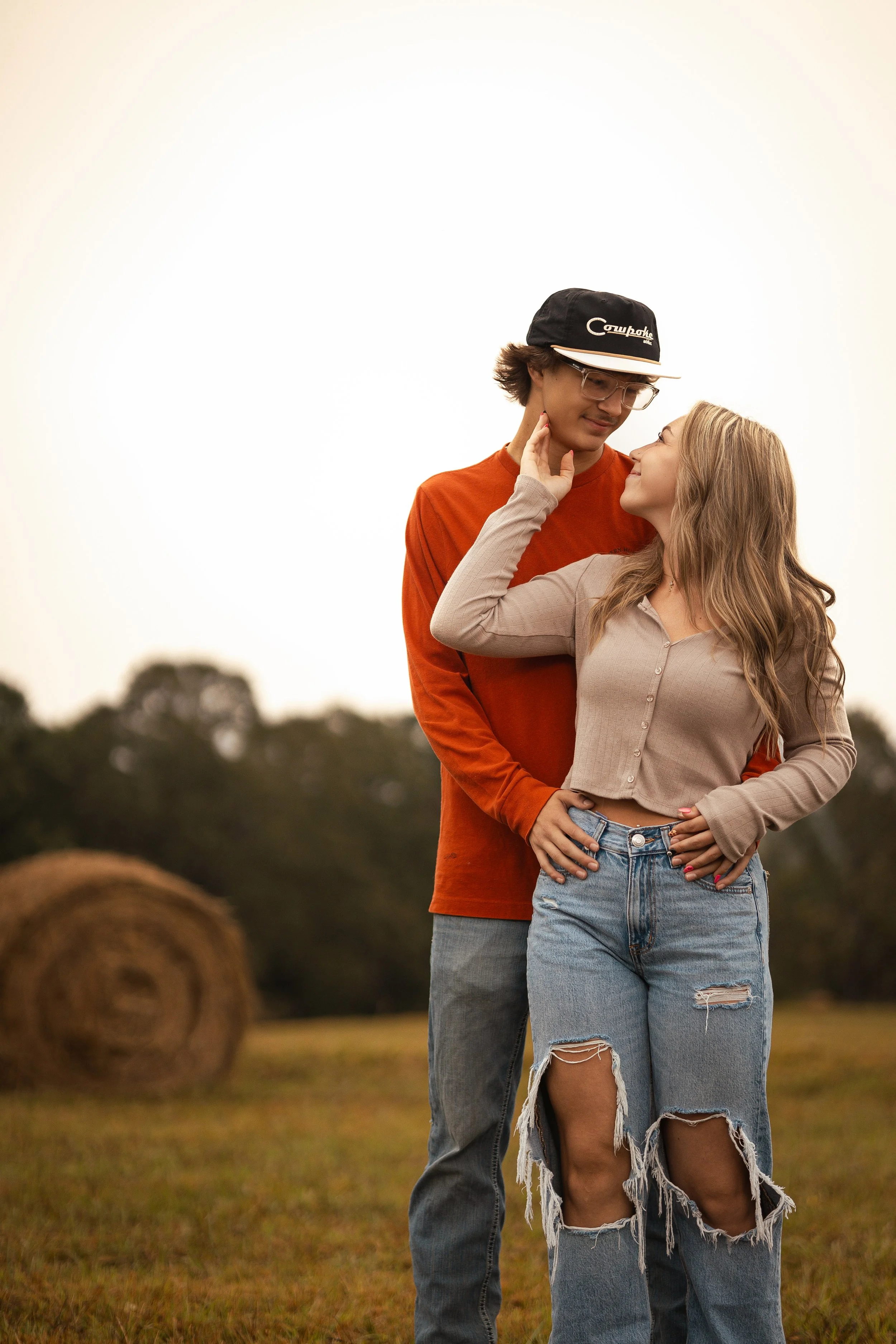 A young couple sharing a tender moment outdoors with hay bales and trees in the background during what appears to be a sunset.