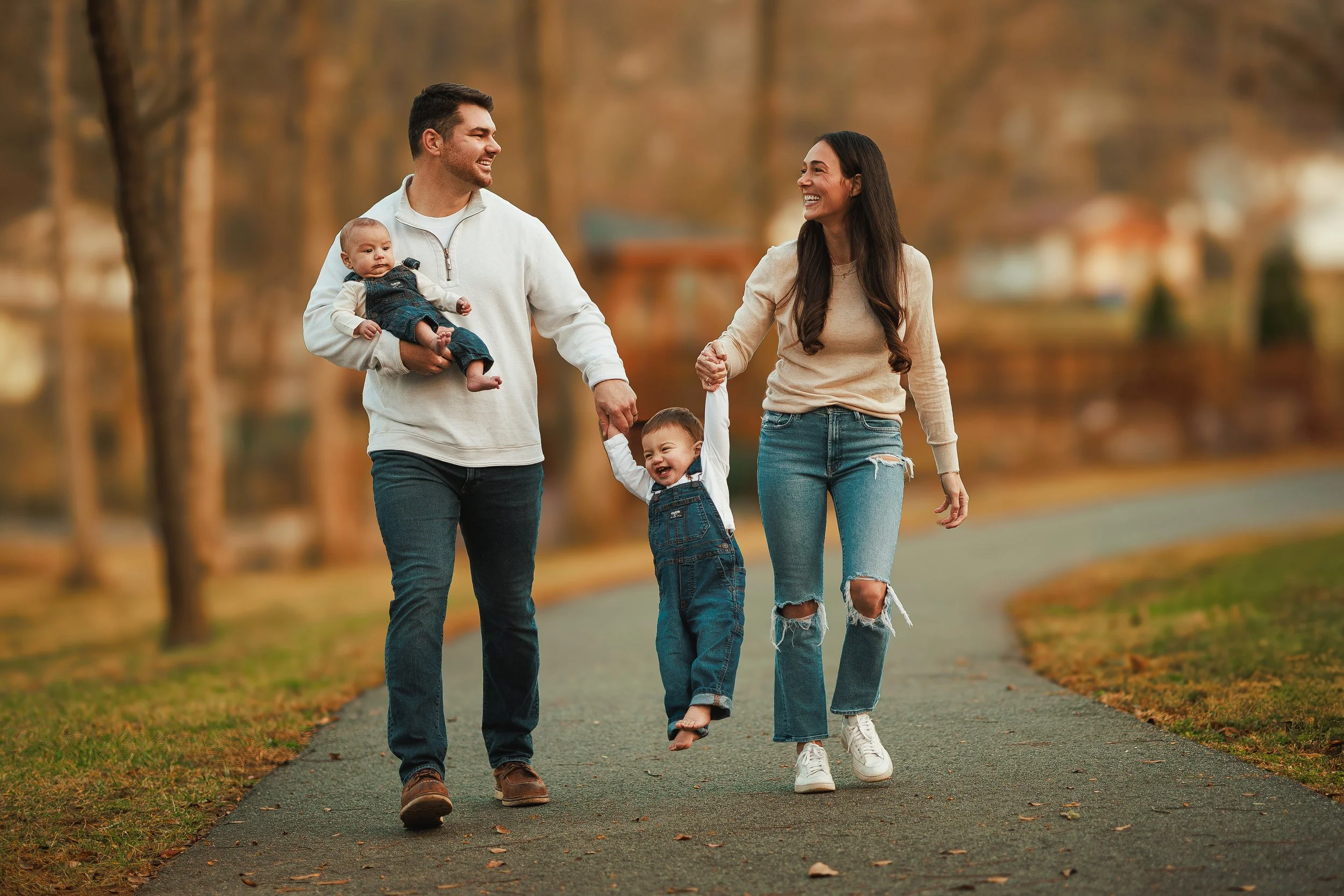 A happy family of four walking on a pathway in a park during fall, holding hands and smiling. The father is carrying a baby, and the mother is holding the hand of a young girl.