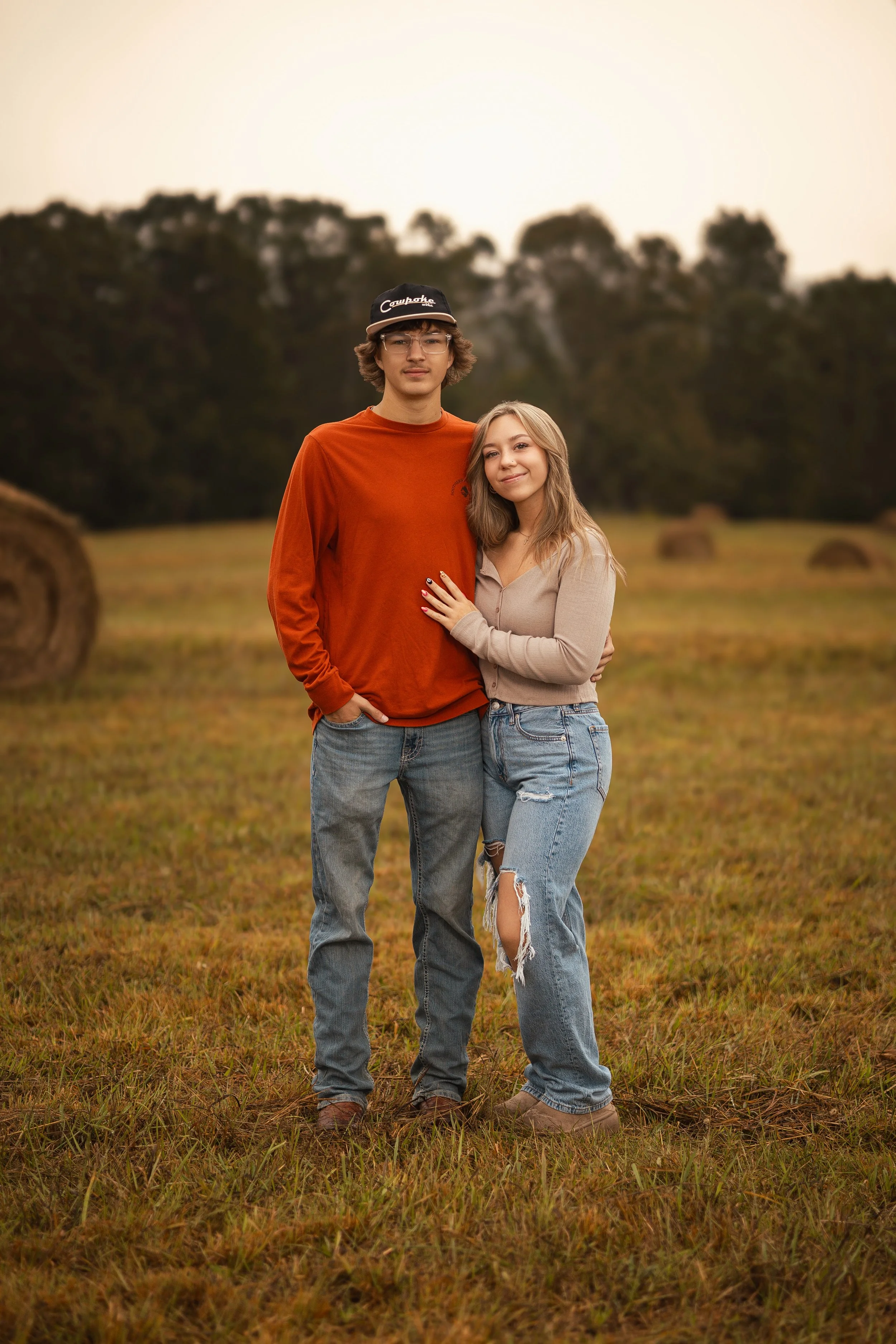 A young couple standing outdoors in a field with hay bales, with trees in the background during sunset, posing closely together with the woman smiling and the man with a neutral expression.