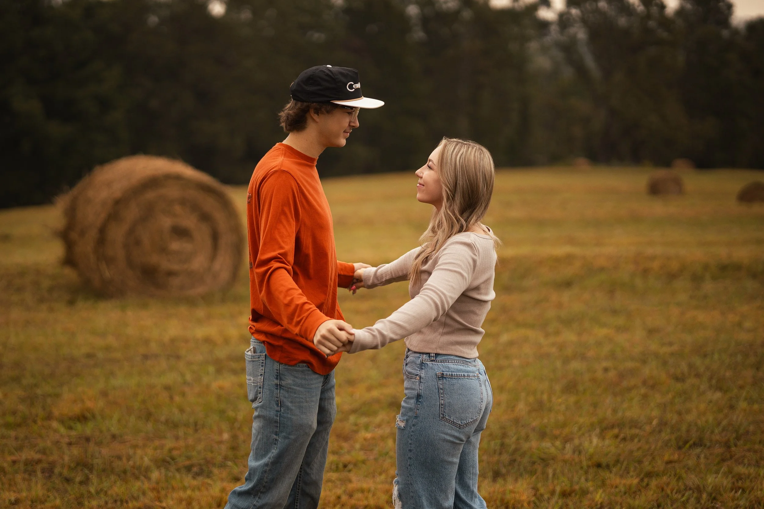 A young man and woman are holding hands and looking at each other in a grassy field with hay bales and trees in the background.