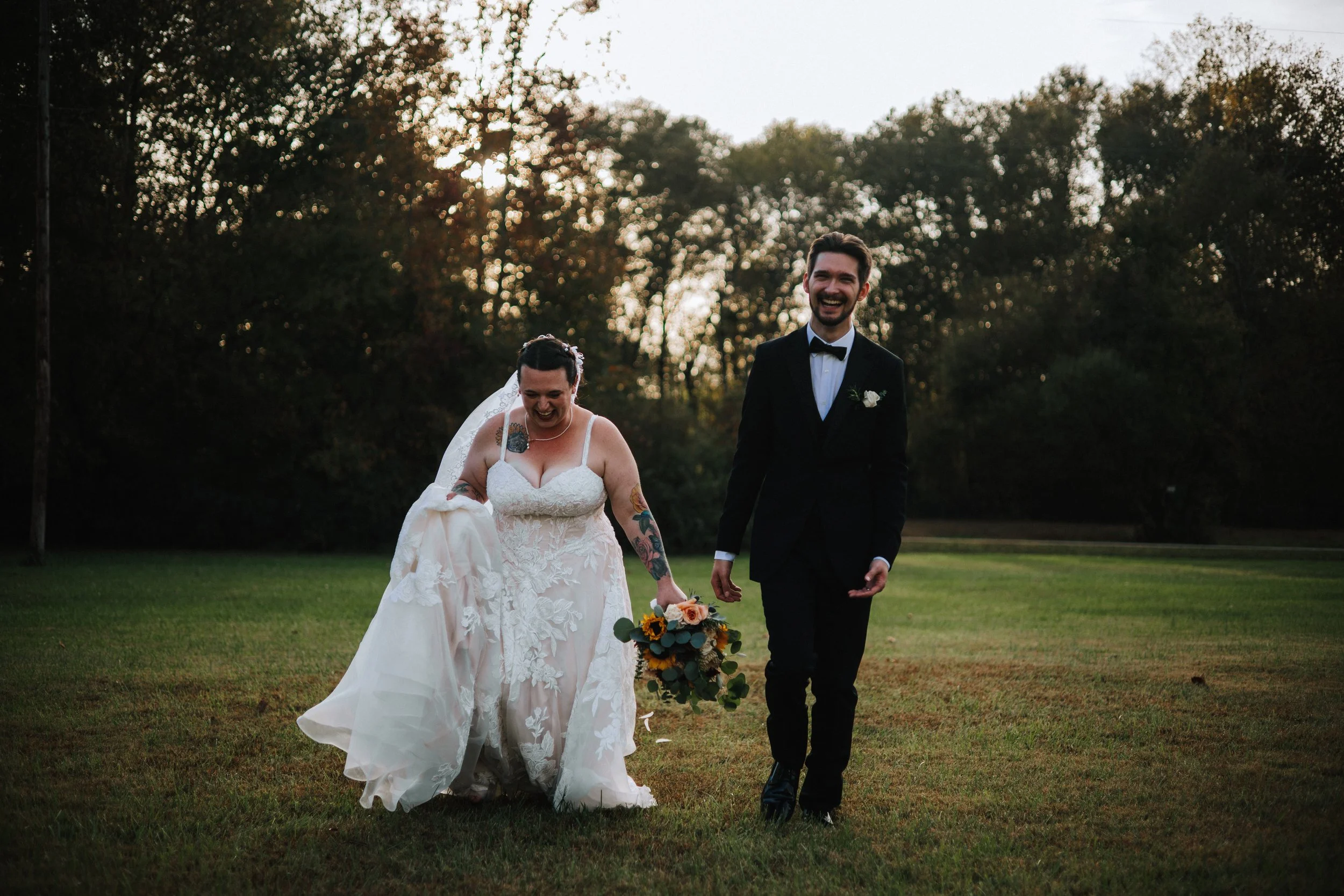 A bride and groom smiling and walking hand in hand on a grassy field during sunset, with trees in the background.