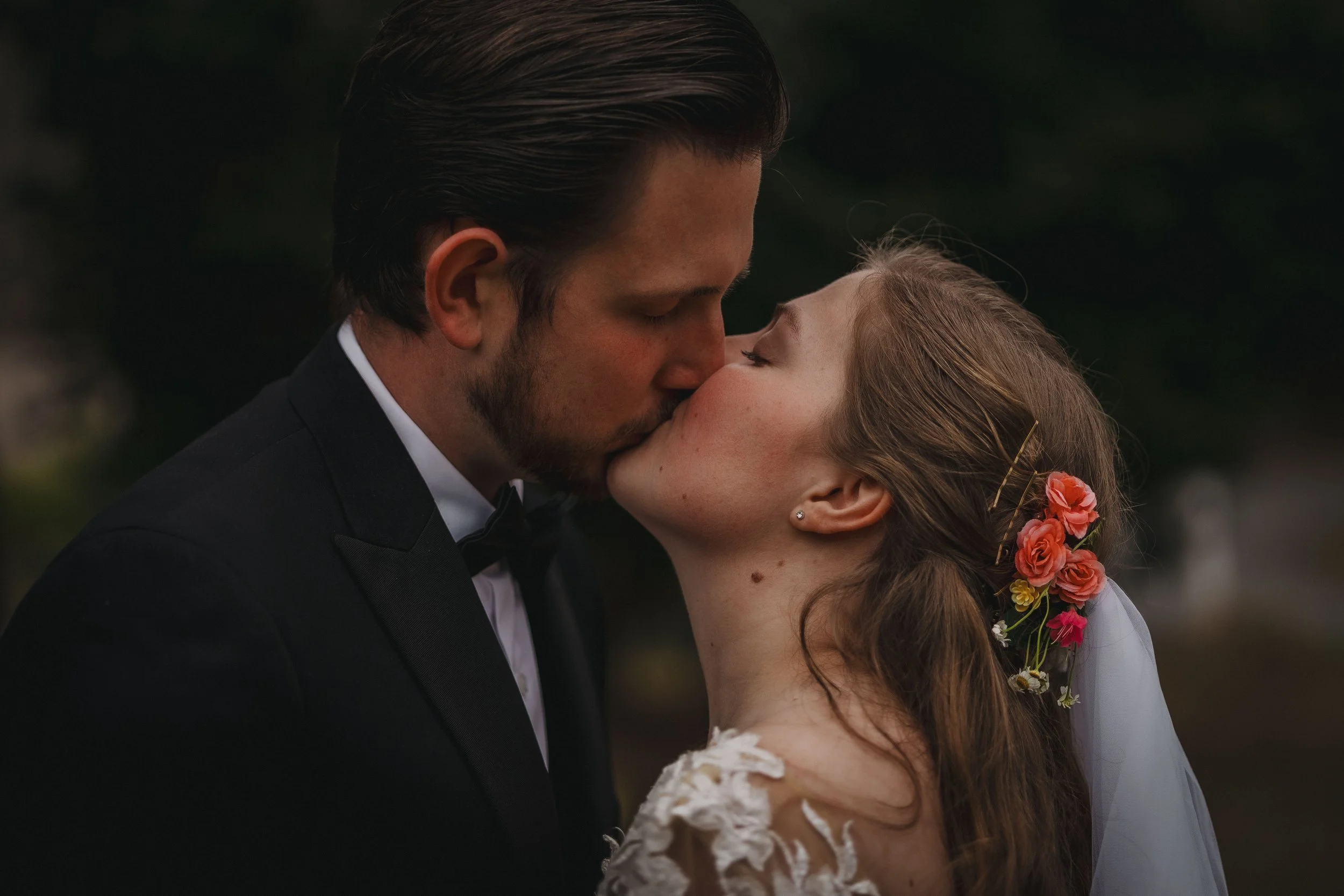 A couple, dressed in wedding attire, sharing a kiss outdoors at dusk, with a dark background.