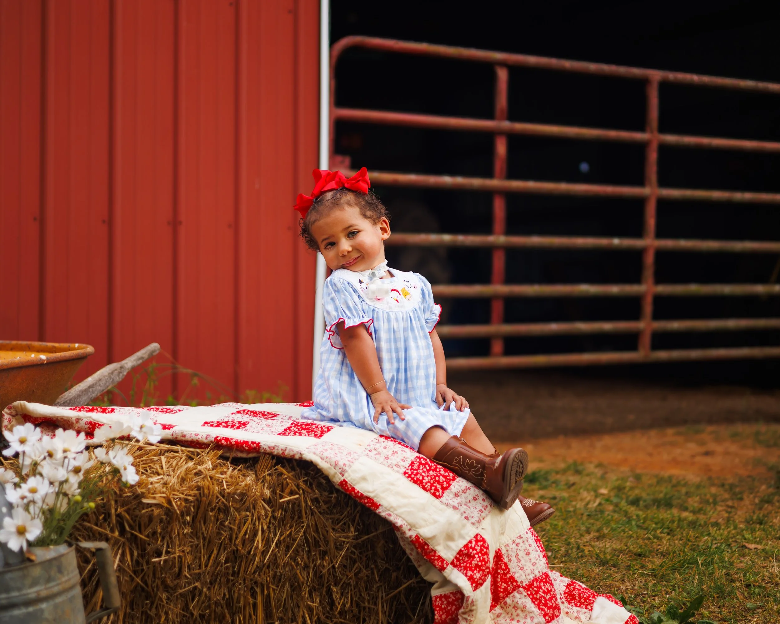 A young girl with curly hair wearing a blue and white checkered dress with smiling face embroidery, a red bow headband, and brown boots, sitting on a quilted blanket on a hay bale in front of a red barn at night.