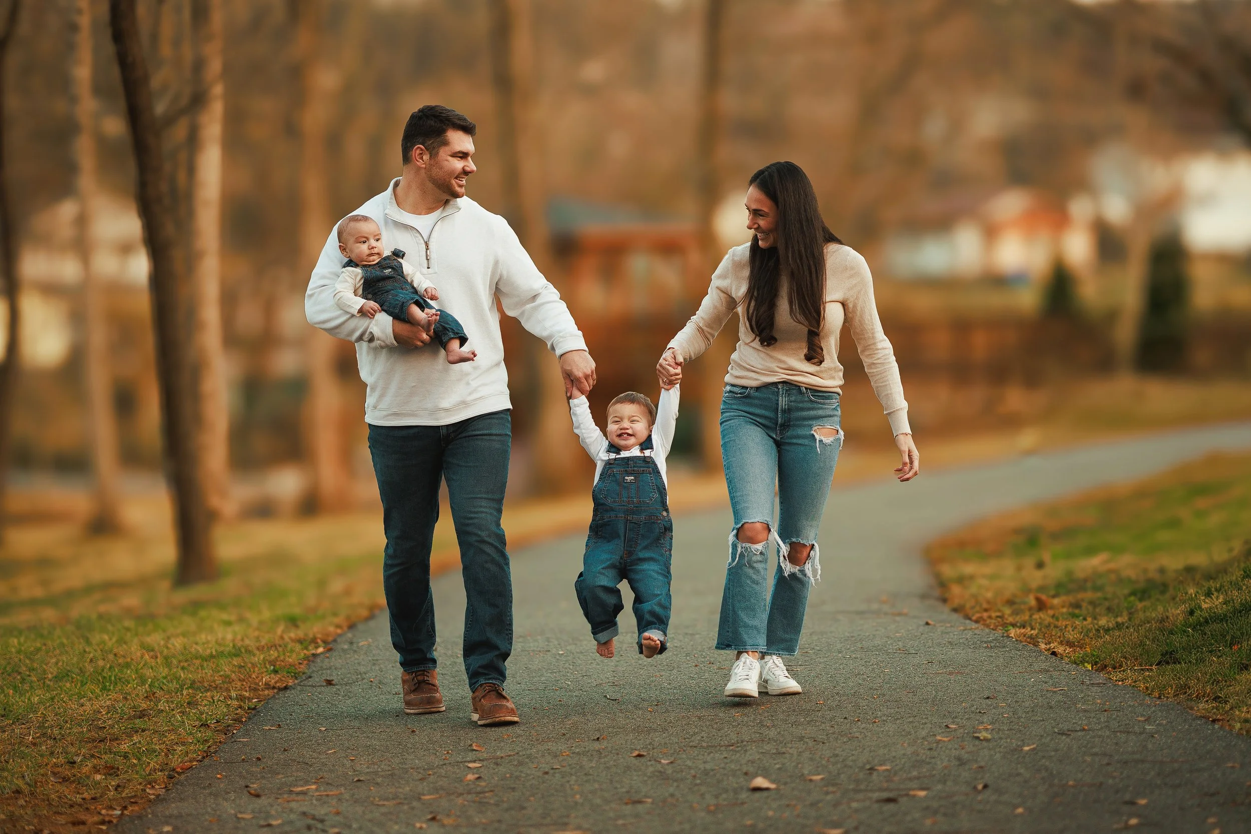 A family of four walking together on a paved path in a park during fall. The baby is being carried by the father, who is holding the child's hand. The mother, wearing ripped jeans and white sneakers, is holding the toddler's other hand as they smile 