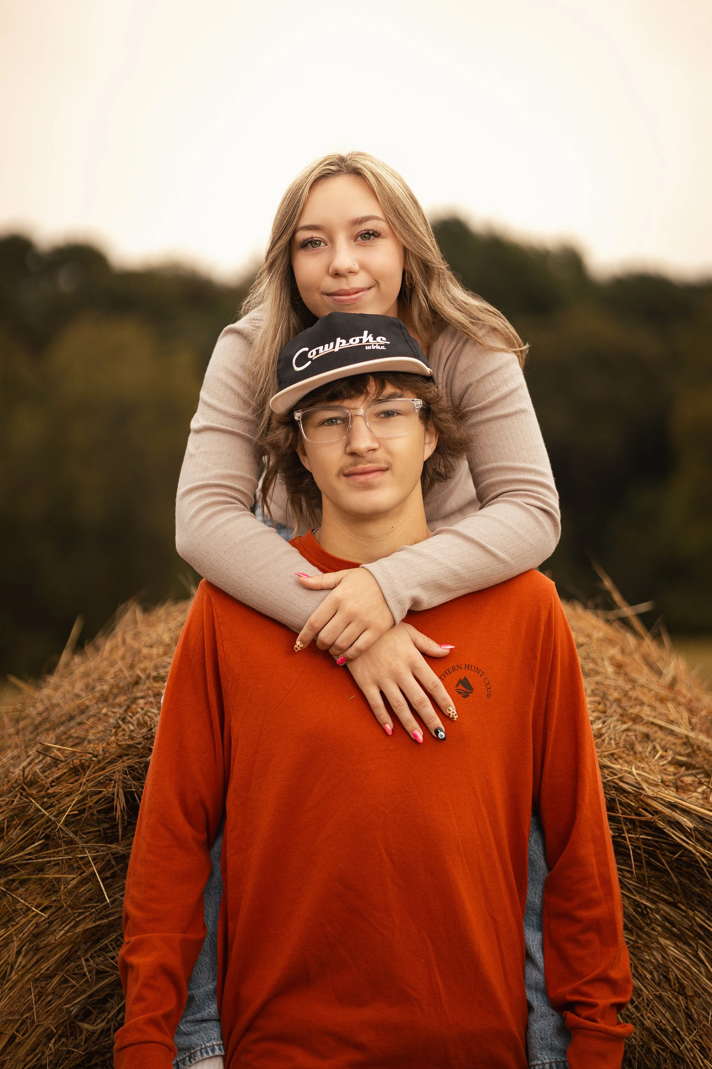 A young woman with long blonde hair leaning on a young man with glasses and curly hair, who is wearing a black cap that says 'Cawpaha' and an orange t-shirt with a logo on it, sitting on a pile of hay outdoors during golden hour.