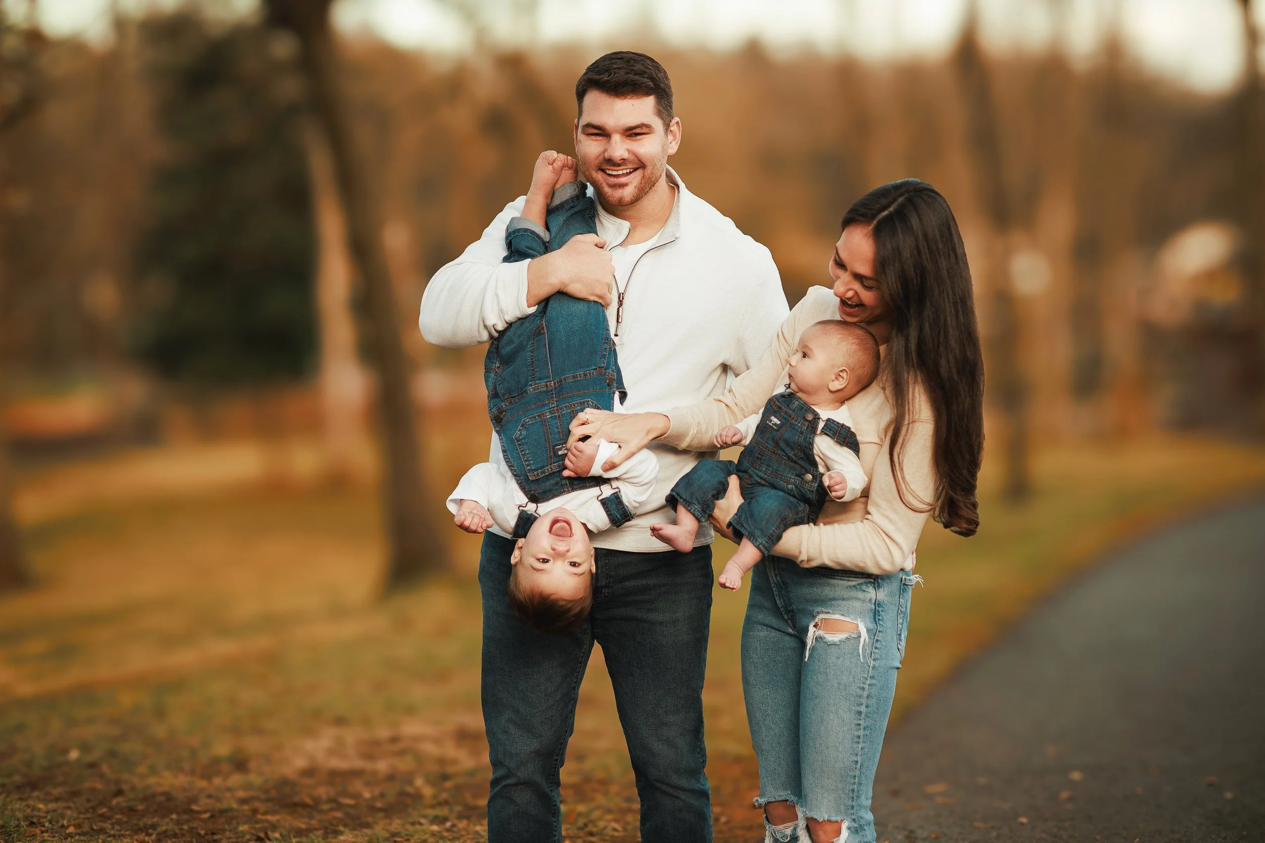 A happy family of four enjoying a walk outdoors in a park with autumn foliage. The father is holding a young boy upside down while the mother is holding a baby girl.