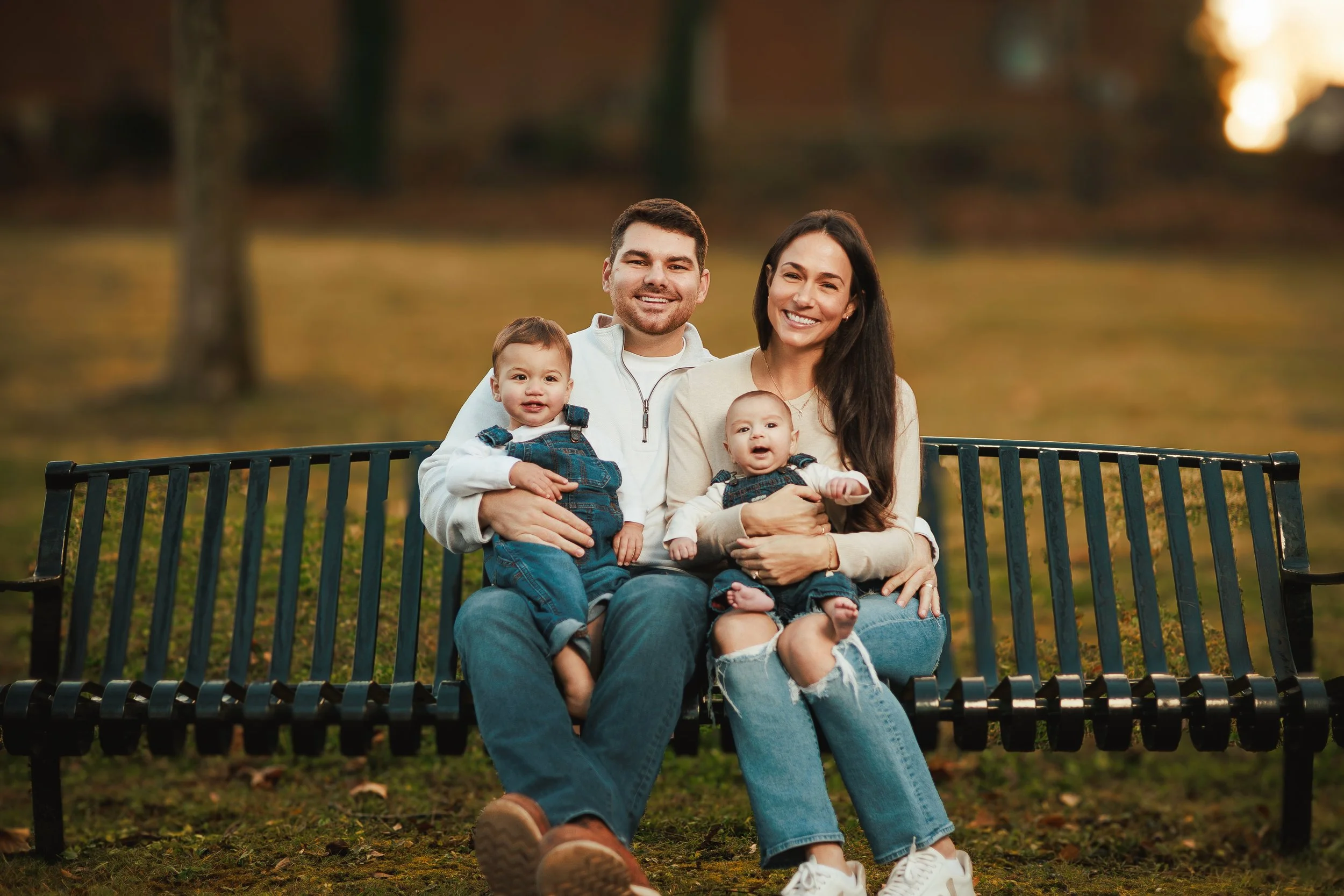 A family of four sitting on a park bench outdoors during autumn. The family consists of a man, woman, and two young children. They are smiling and enjoying the moment. The background shows blurred trees with fall foliage.