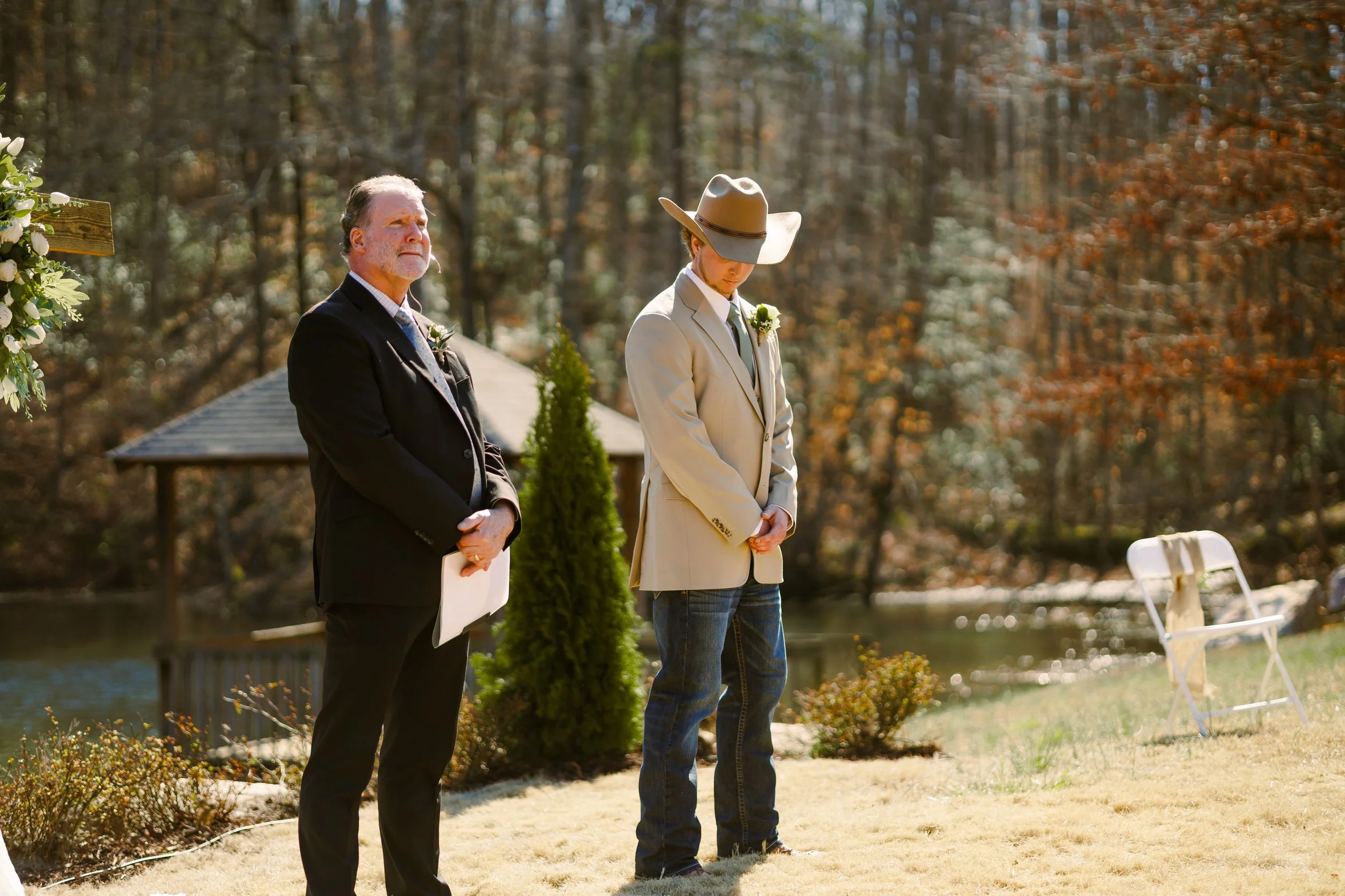 A man in a dark suit and an elderly man in casual attire stand outdoors during a ceremony, likely a wedding, in a park with trees and a small body of water in the background.