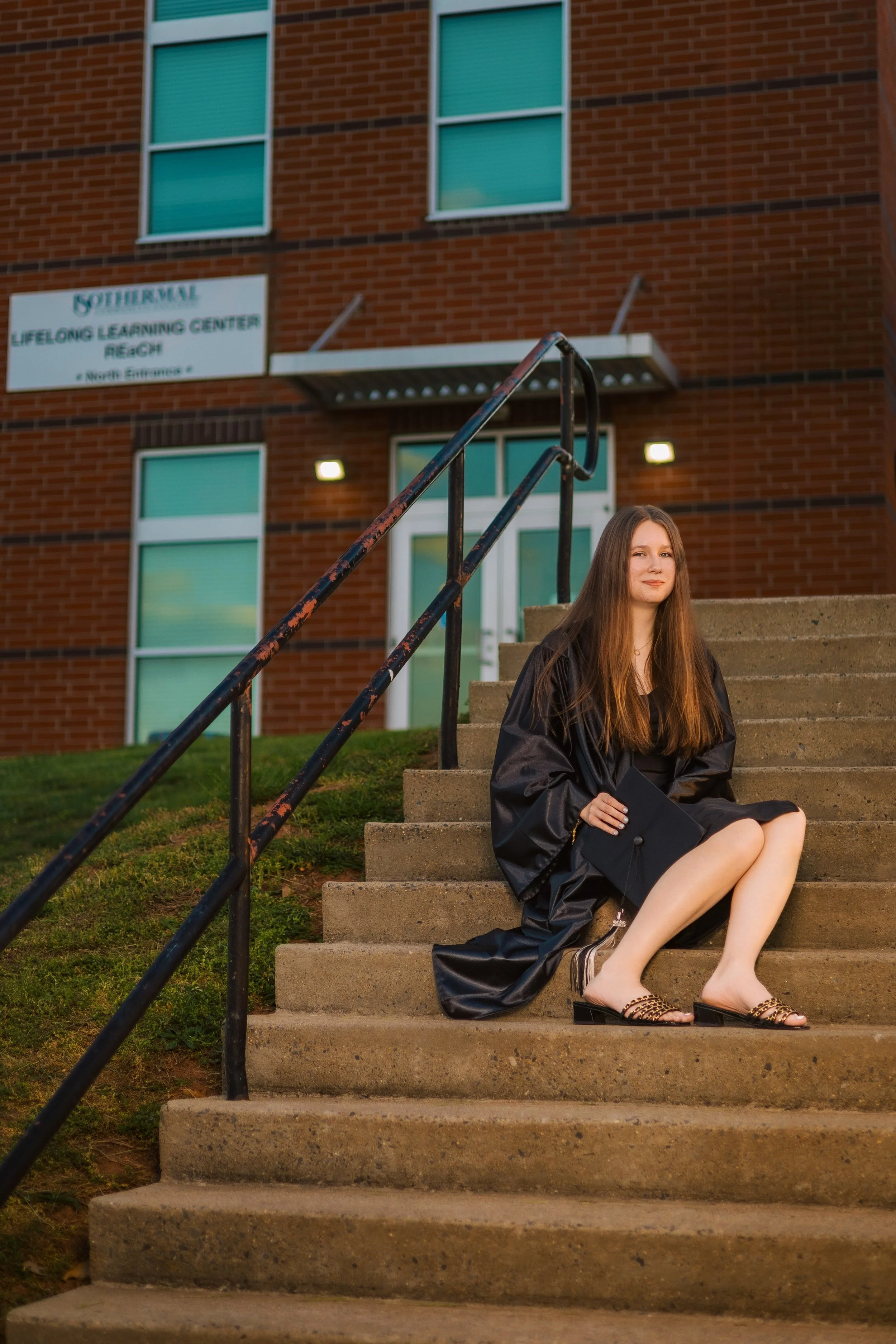 A young woman in a black graduation gown sitting on outdoor concrete stairs, holding a diploma, with a brick building and a sign for a lifelong learning center in the background.