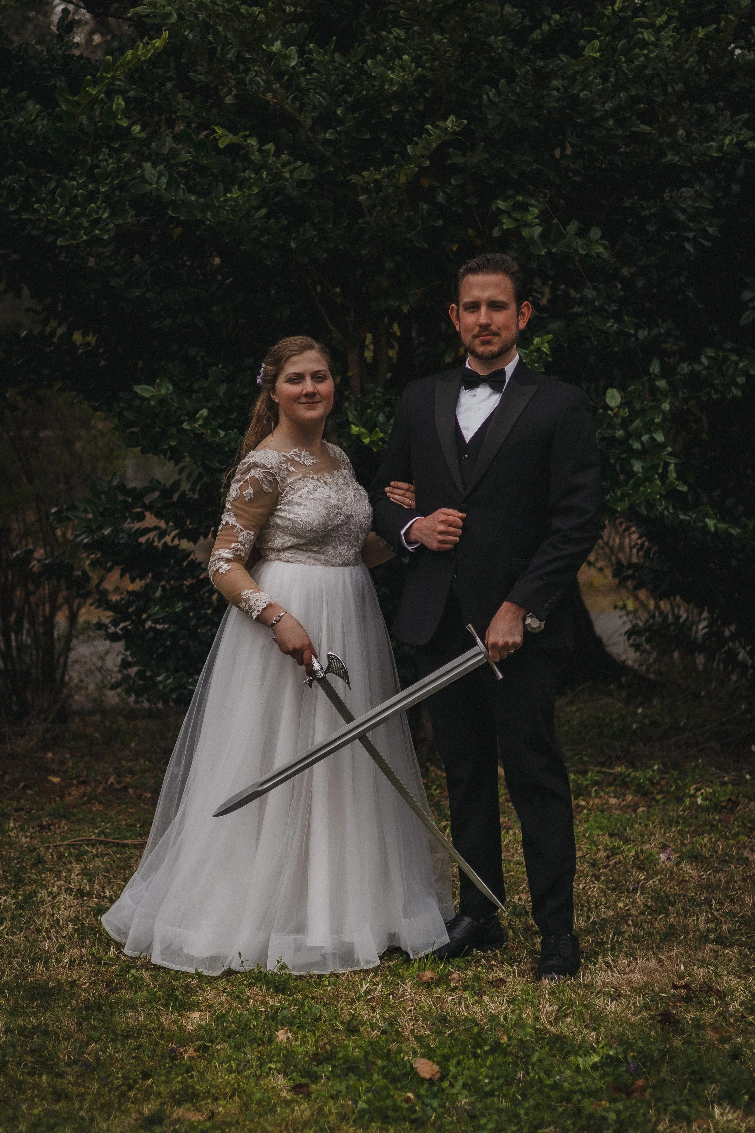 A woman in a wedding dress and a man in a black tuxedo pose outdoors, holding swords, with a large leafy tree in the background.