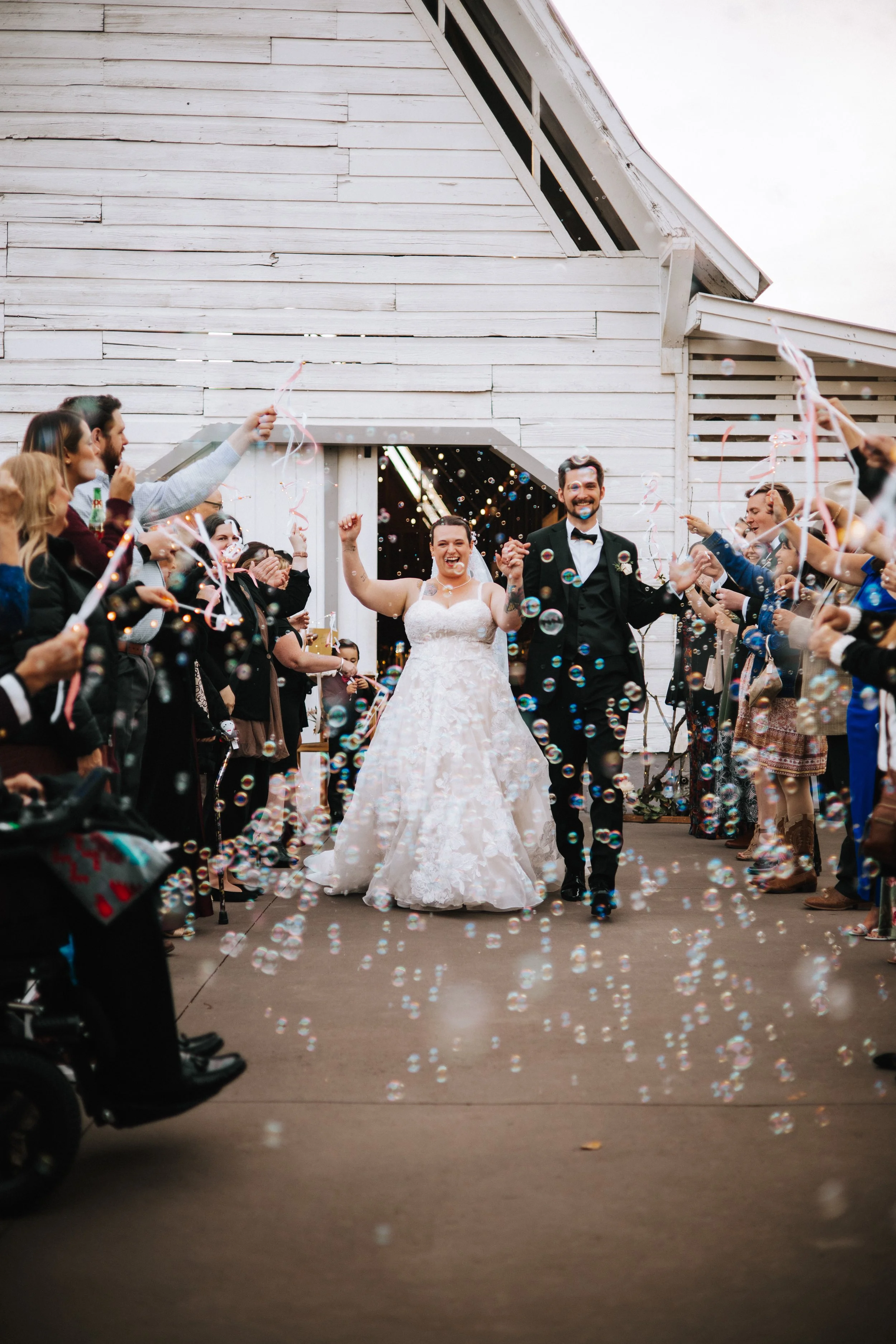 A newly married couple walking out of a barn-style venue after their wedding reception, surrounded by cheering friends and guests, with bubbles and streamers in the air.