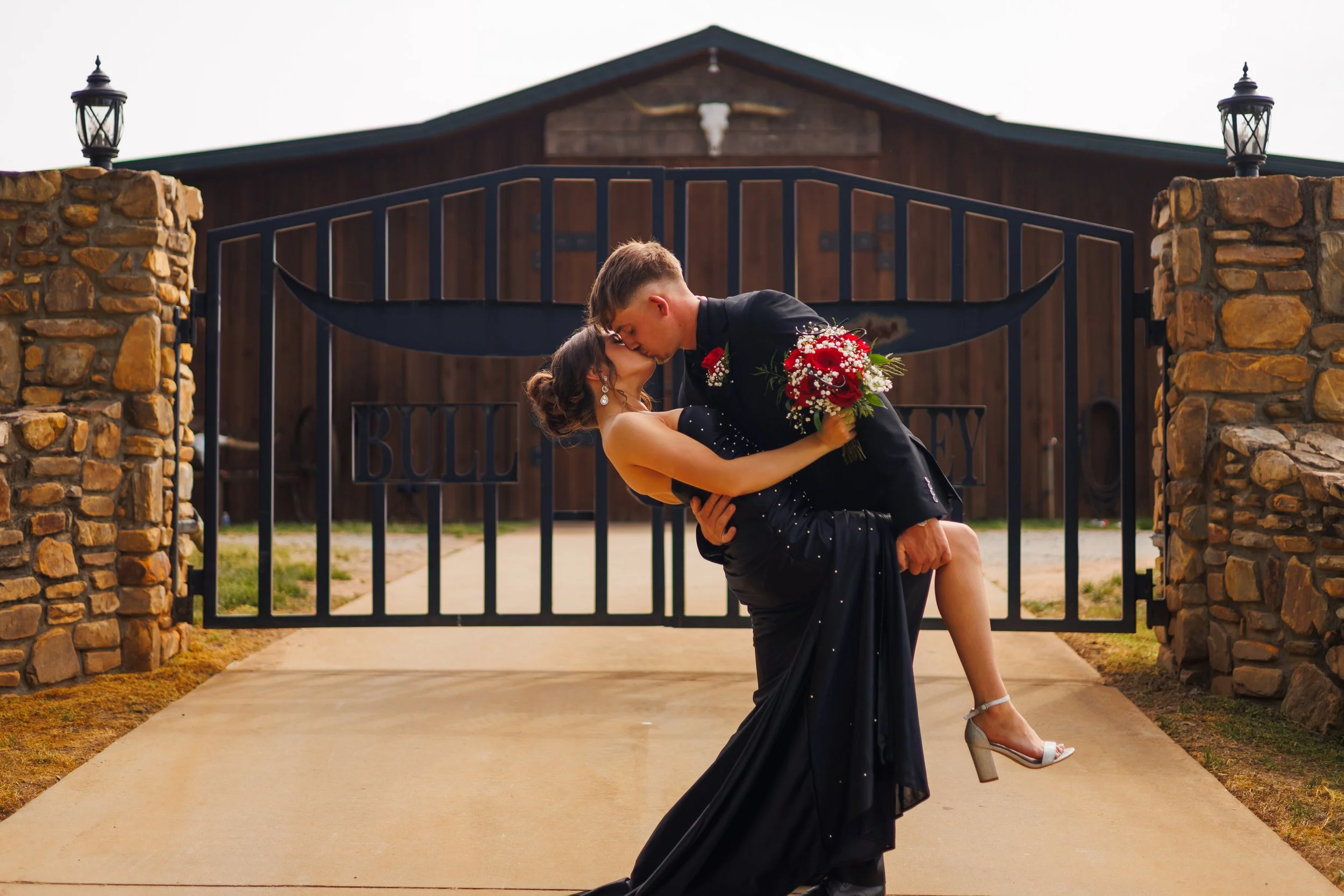 A couple embracing and kissing in front of a gated entrance to a building, with stone pillars and lanterns on top of the pillars.