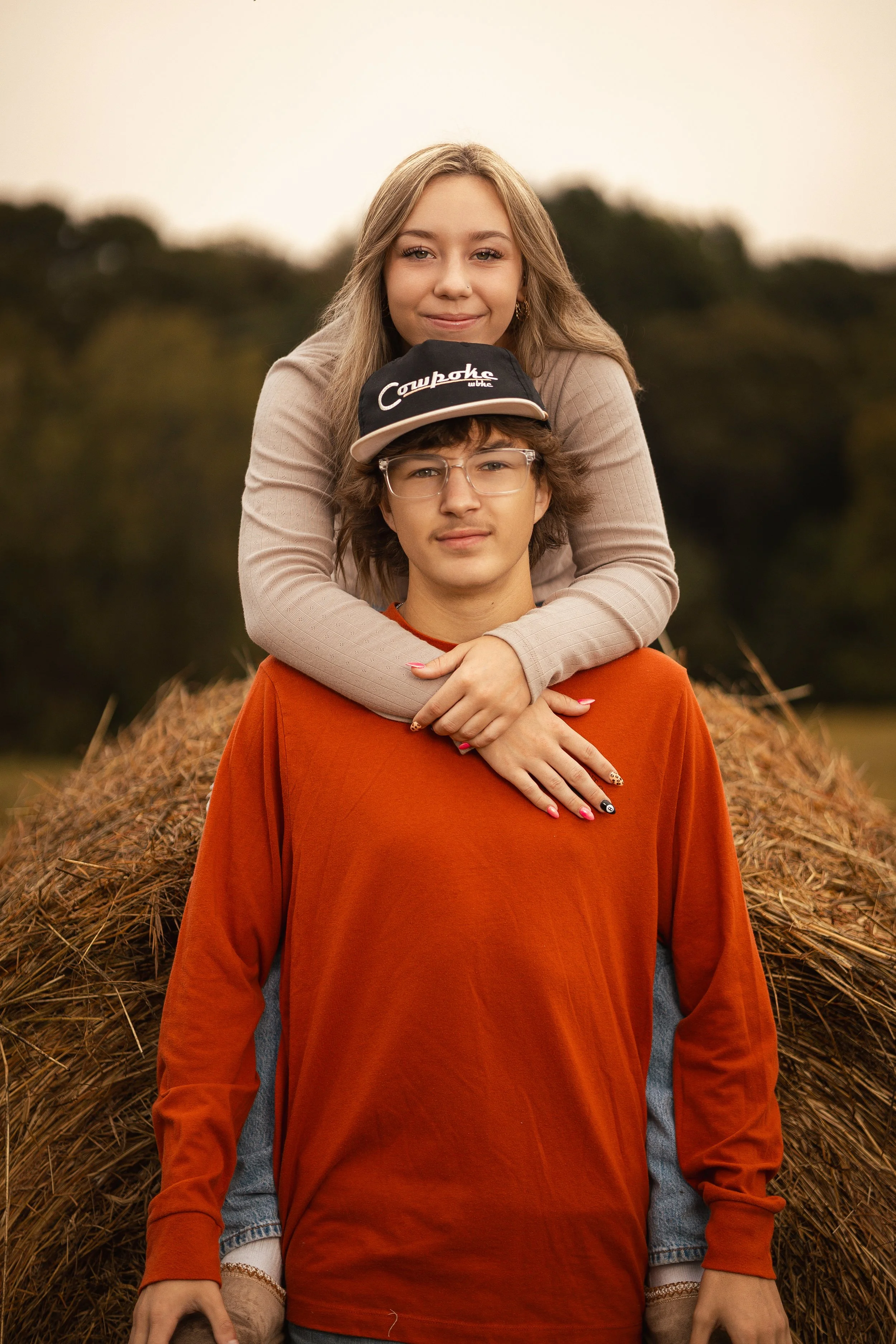 A young girl with long blonde hair sitting behind a young man with glasses and a cap that says 'Cowboys'. She is smiling, and they are outdoors in front of a pile of hay on a cloudy day.
