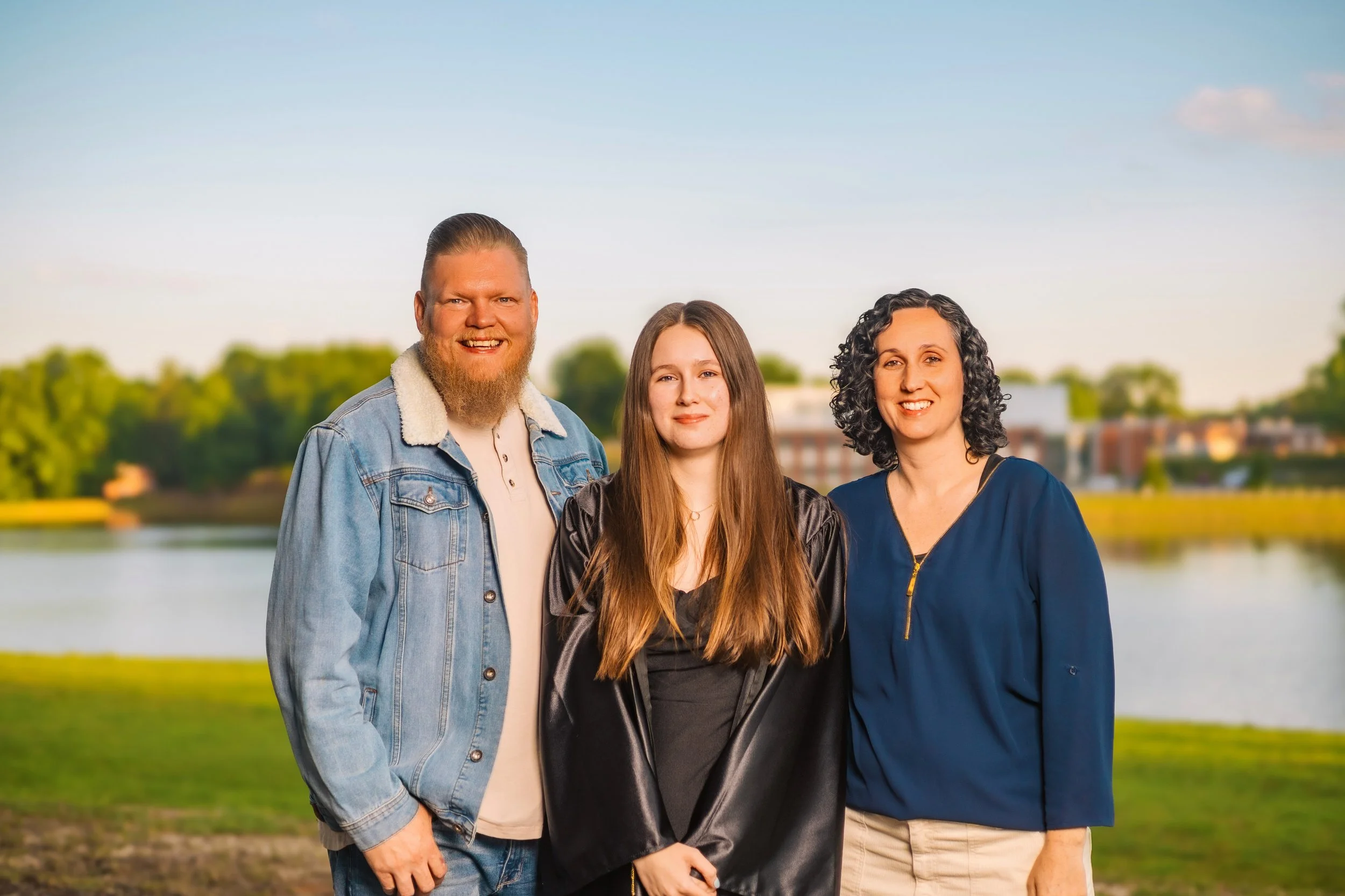 Group of three people standing outdoors near a river or lake, with trees and buildings in the background, during sunset or sunrise.