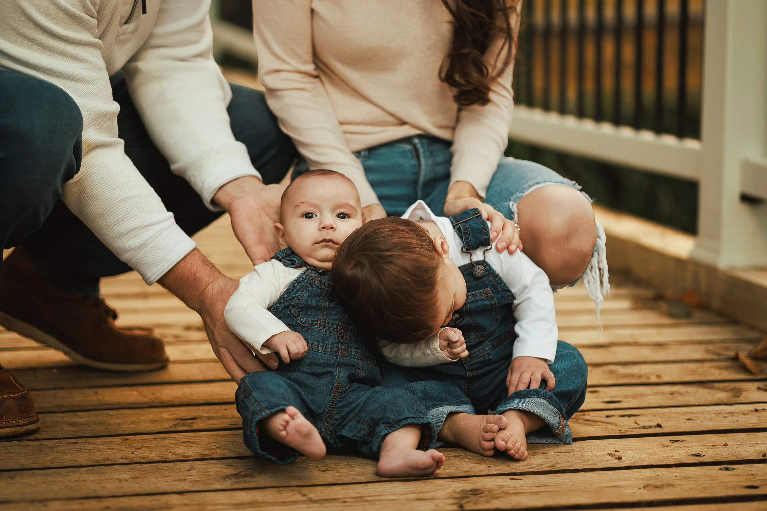 Two children lying on a wooden deck with one looking at the camera and the other with head bowed, while two adults support their heads and arms.