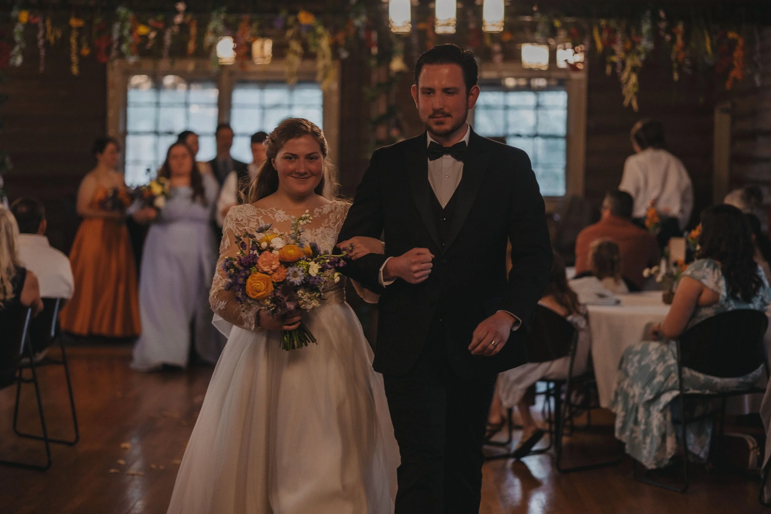 A bride and groom walking down the aisle at their wedding ceremony inside a rustic wooden venue with hanging flowers and guests seated at tables.