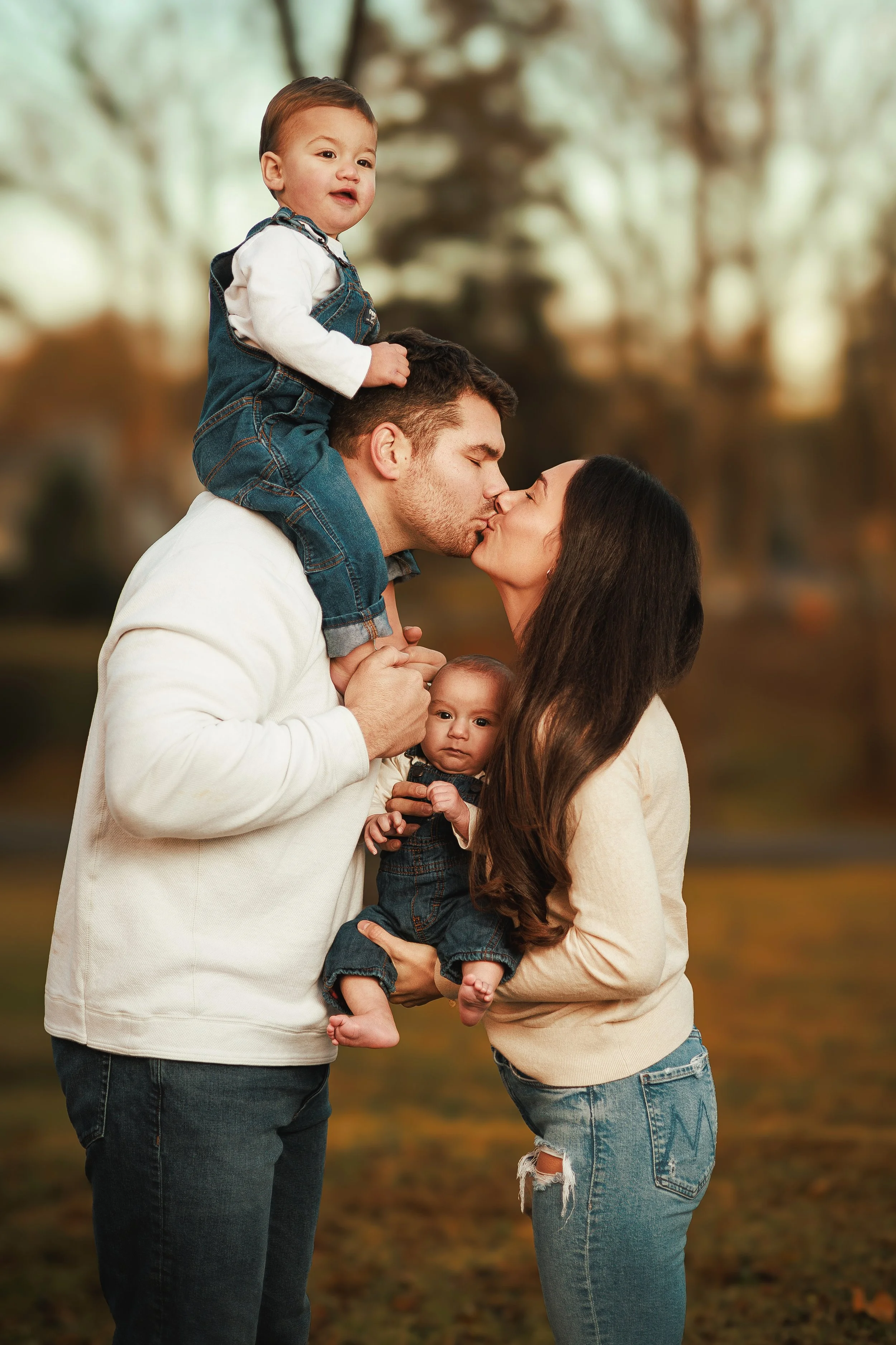 A family of four outdoors in autumn, with a man and woman kissing, the man carrying a young boy on his shoulders and the woman holding a baby girl.